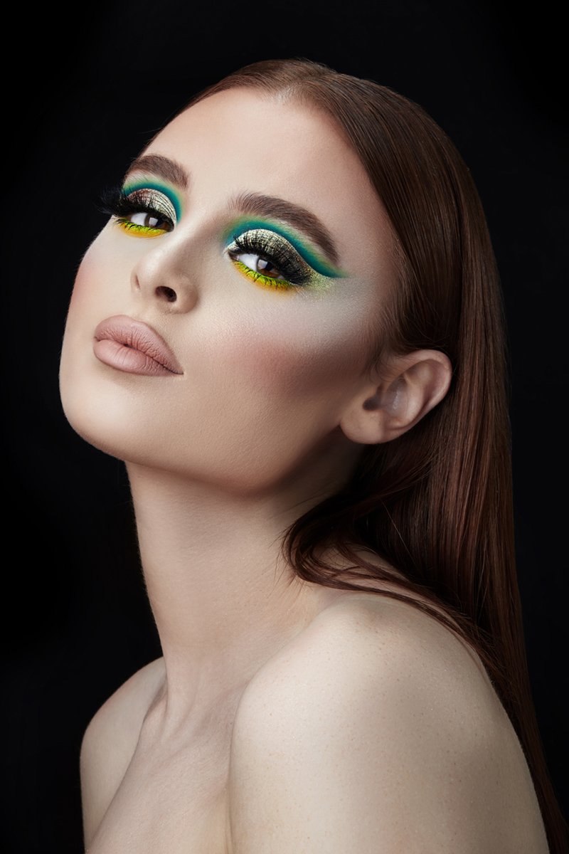 A woman with elaborate eye makeup featuring green and yellow colors, dark eyeliner, and long eyelashes, with natural nude lipstick and straight, auburn hair against a black background.