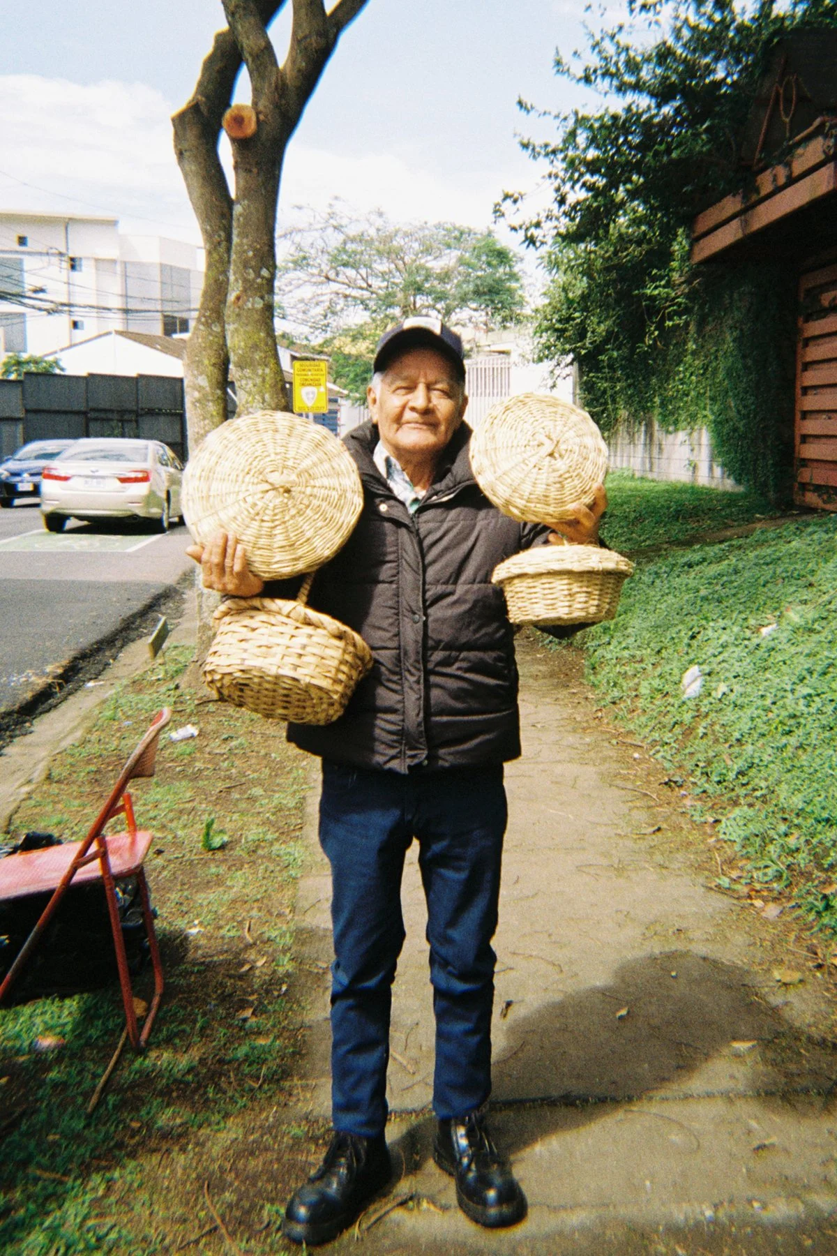An elderly man standing on a sidewalk holding woven baskets in both hands, wearing a black jacket, dark pants, black boots, and a baseball cap, with cars and trees in the background.