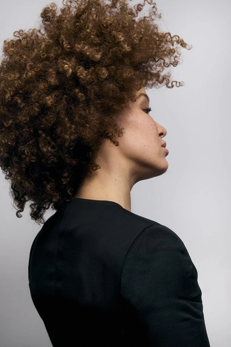 Profile side view of a woman with curly, voluminous brown hair wearing a black top, against a plain light background.
