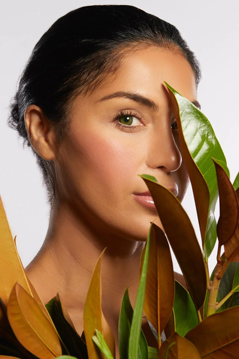 A woman with tan skin and dark hair holding green and brown leaves in front of her face.