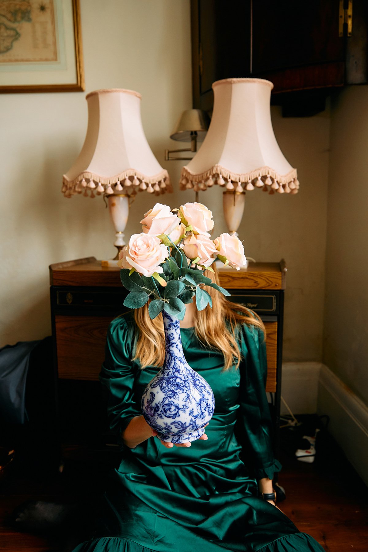 Person sitting in a room holding a blue and white vase with pink roses covering their face, two vintage lamps on a wooden table behind them.