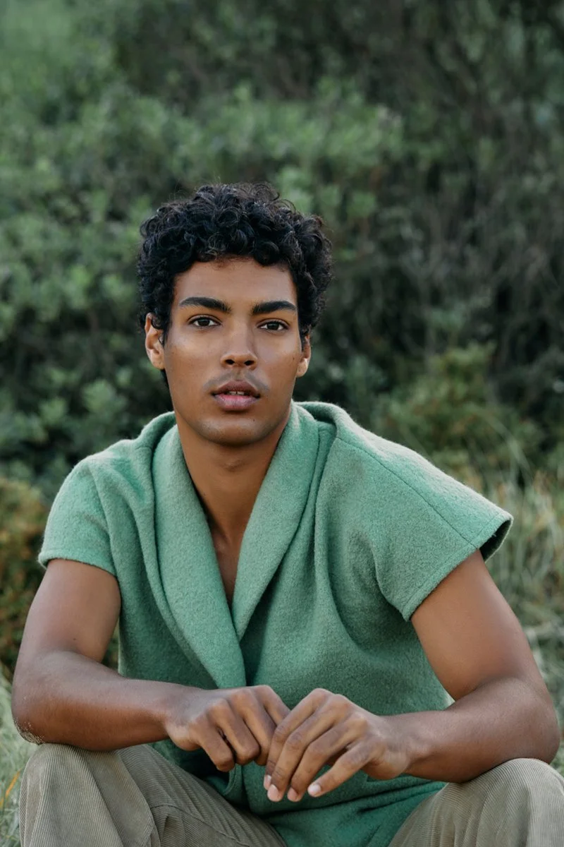 A young man with curly black hair and medium skin sitting outdoors in front of green foliage, wearing a green sleeveless sweater.