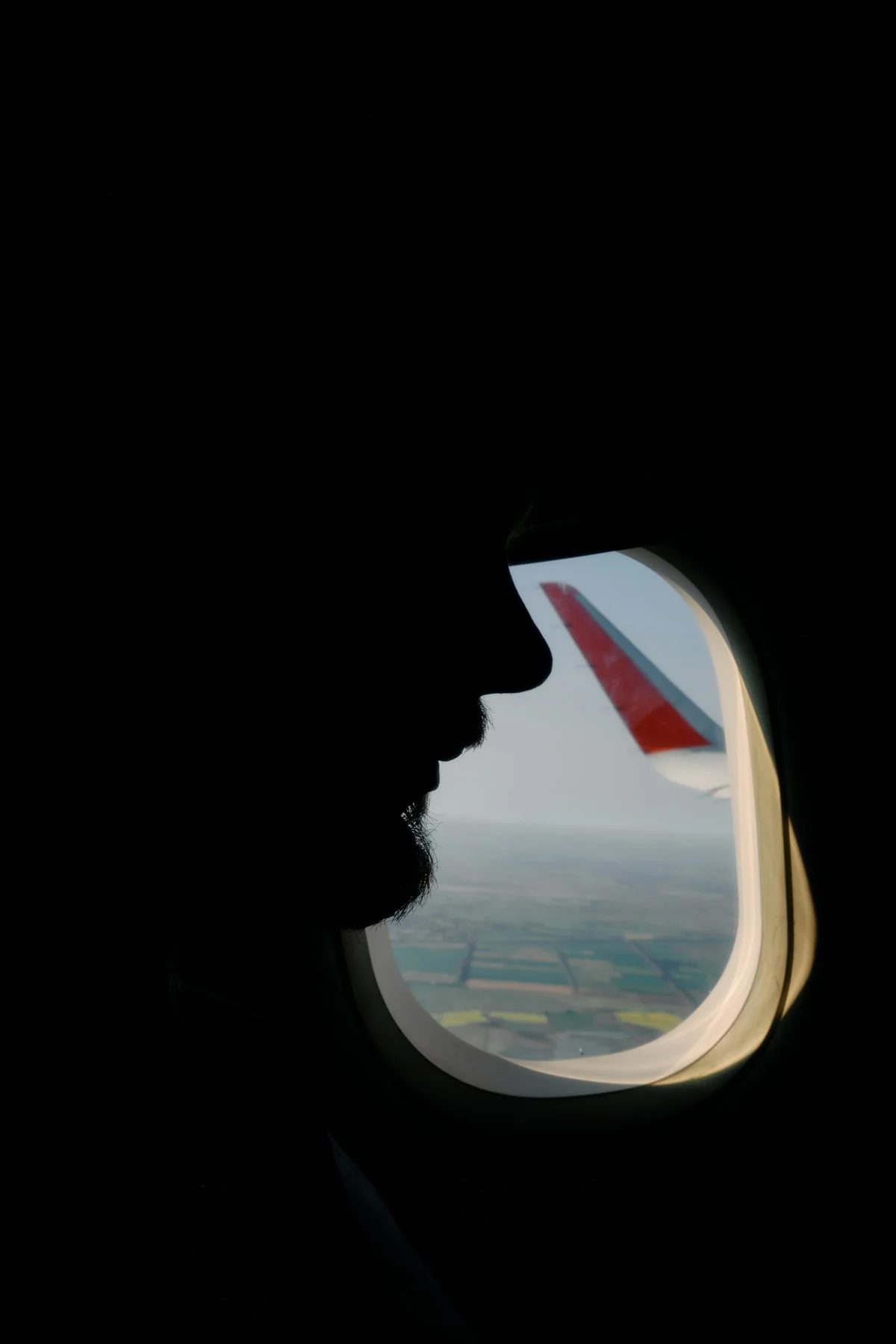 Silhouette of a man with a beard looking out the airplane window at the landscape below, including fields and a red-and-gray airplane wing.