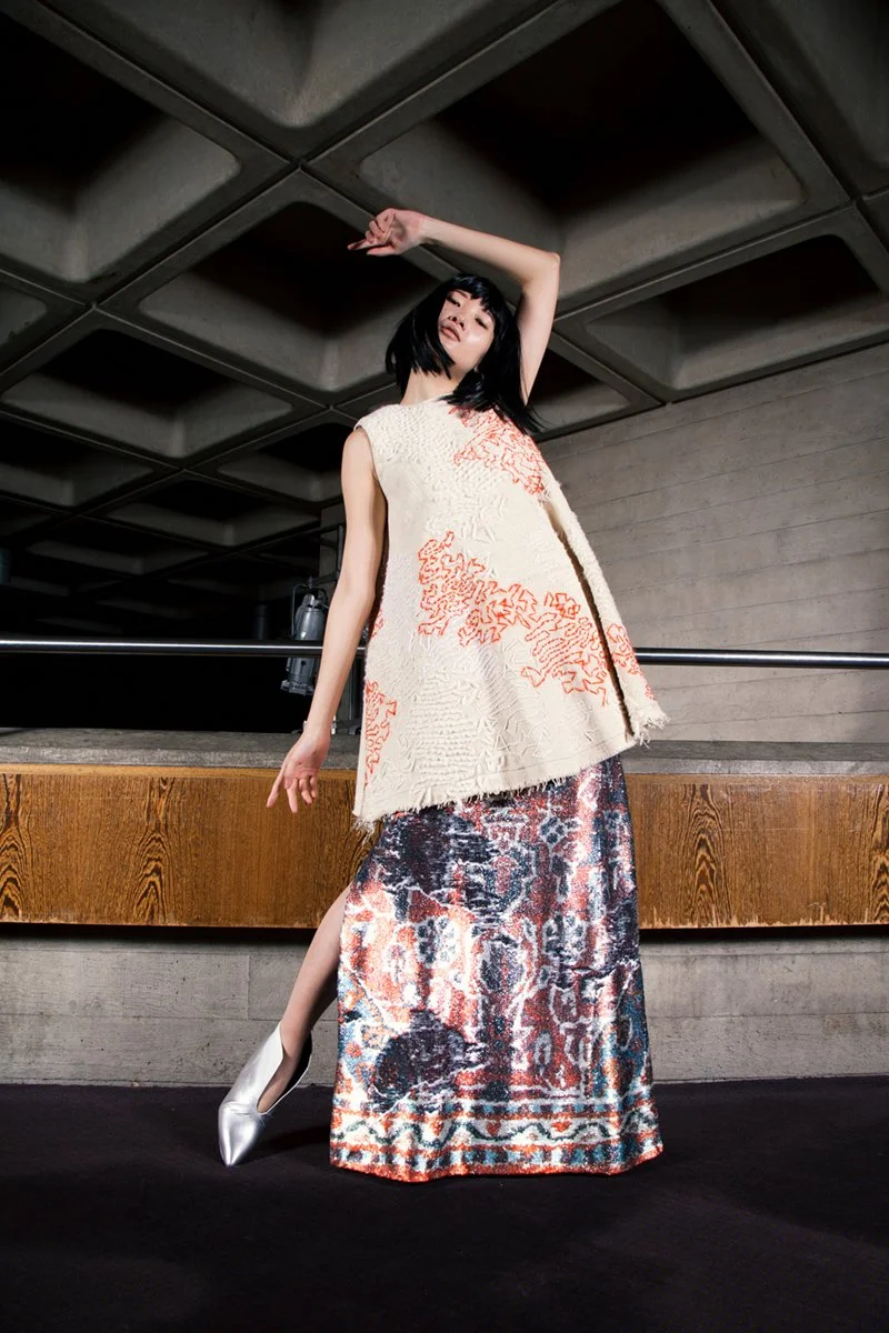 A woman with black hair in a bob haircut, wearing a sleeveless cream-colored top with red embroidery and a long, colorful patterned skirt, standing in a modern interior space with a concrete wall and wooden counter behind her.