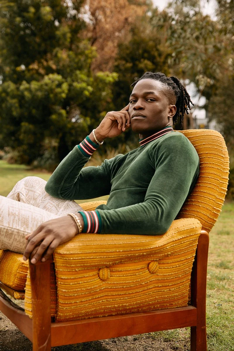 A young man with dreadlocks sitting on a yellow vintage armchair outdoors with trees in the background, wearing a green long-sleeve shirt and patterned pants.