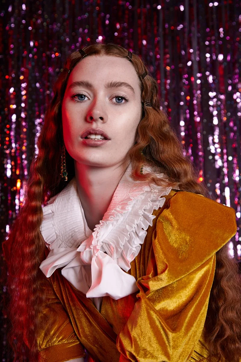 Portrait of a young woman with long, wavy red hair and blue eyes, wearing a mustard yellow velvet jacket with a white ruffled collar and a large bow, standing in front of a purple sequin backdrop.