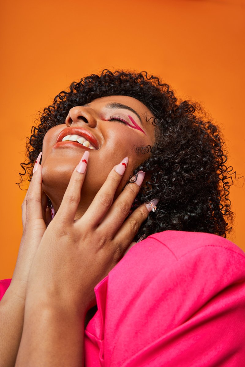 A woman with curly hair smiling with her eyes closed, wearing pink and with colorful makeup, against an orange background.
