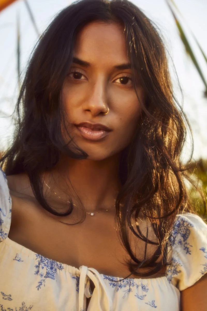 Close-up portrait of a young woman with dark hair, wearing a white blouse with blue embroidery, and a delicate necklace, outdoors at sunset.