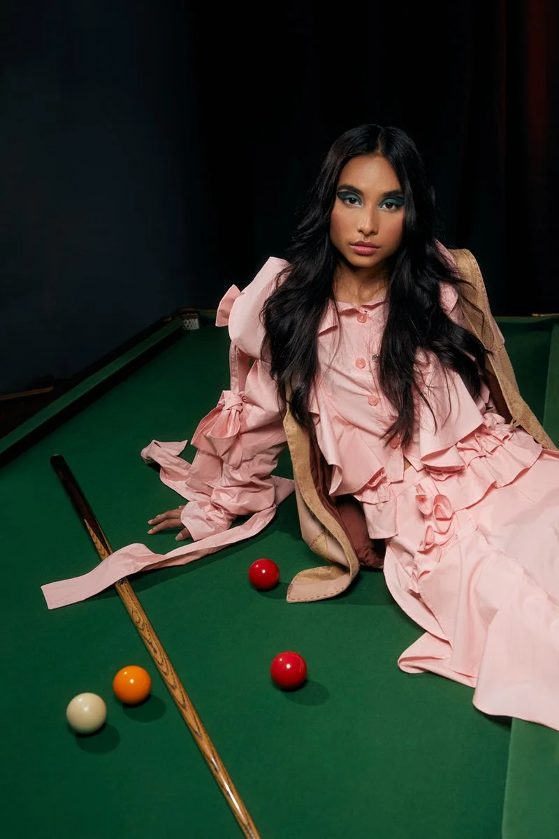 A woman dressed in a pink dress with ruffles and blue eyeshadow, lounging on a green pool table with three colored balls and a cue stick.