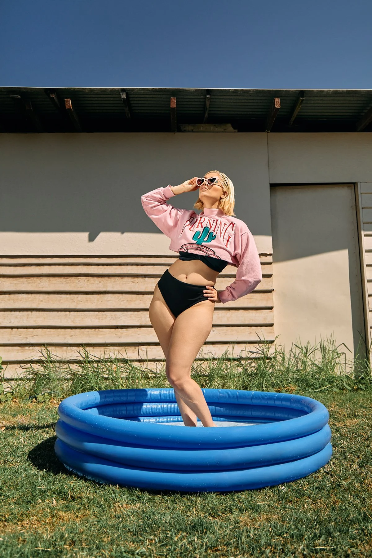 A woman stands in an inflatable pool outdoors, wearing sunglasses, a pink sweatshirt, and black swim bottoms. She is holding her sunglasses and looking up, with a background of a modern building and clear blue sky.