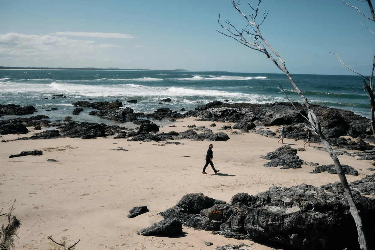 People walking on a rocky and sandy beach with ocean waves and a cloudy sky.