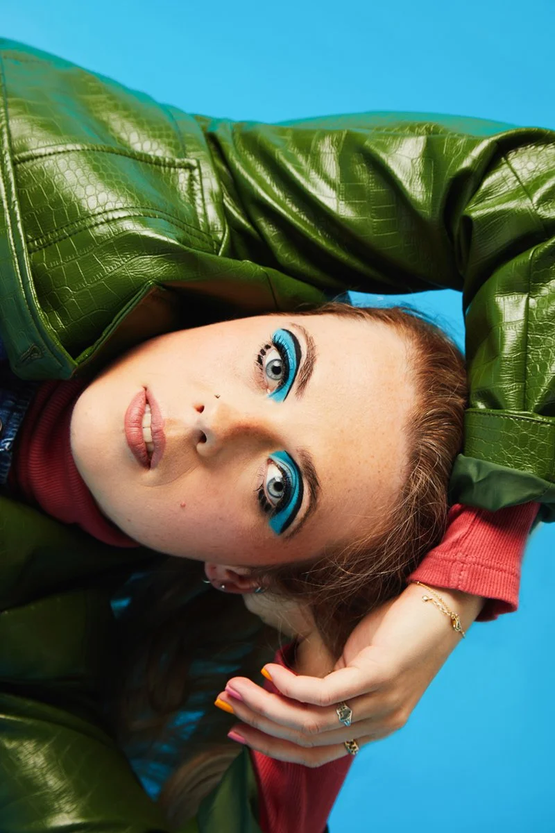 A woman with vibrant blue eye makeup, wearing a green leather jacket and red shirt, lying against a bright blue background, with her hand resting on her head.