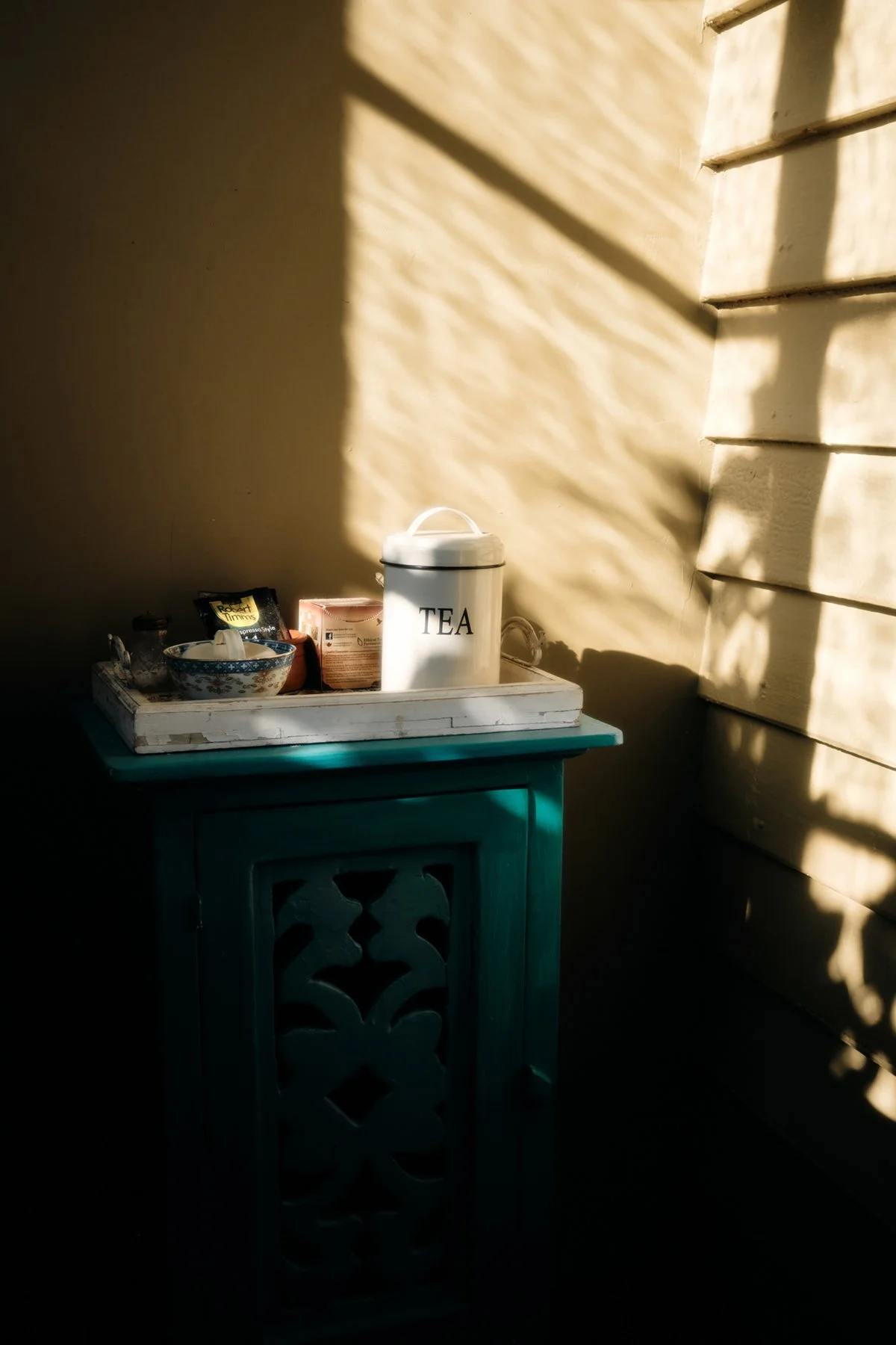 A small teal wooden table with a white tray on top holds a white container labeled 'TEA', some boxes, and cups. Sunlight casts patterned shadows on the wall behind and to the side of the table.