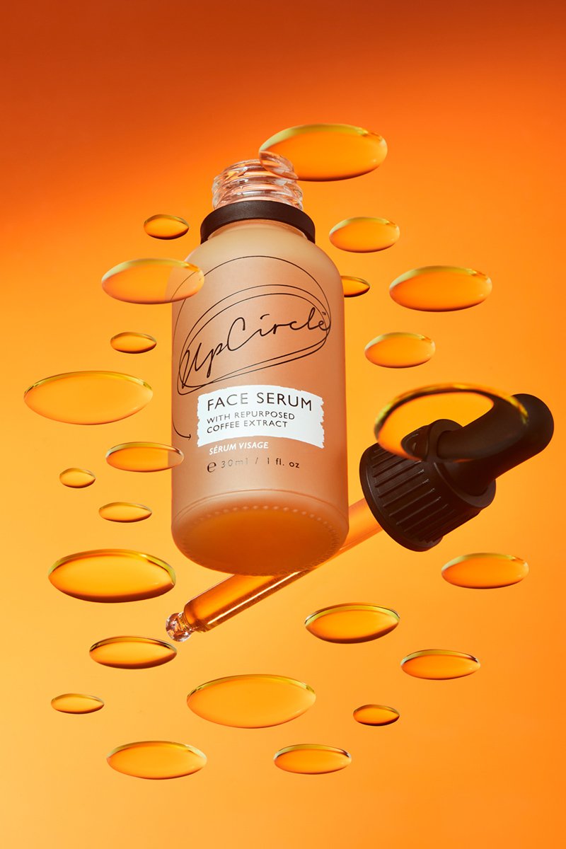 A bottle of Hip Circle face serum with repurposed coffee extract, surrounded by orange and yellow droplets on an orange background.