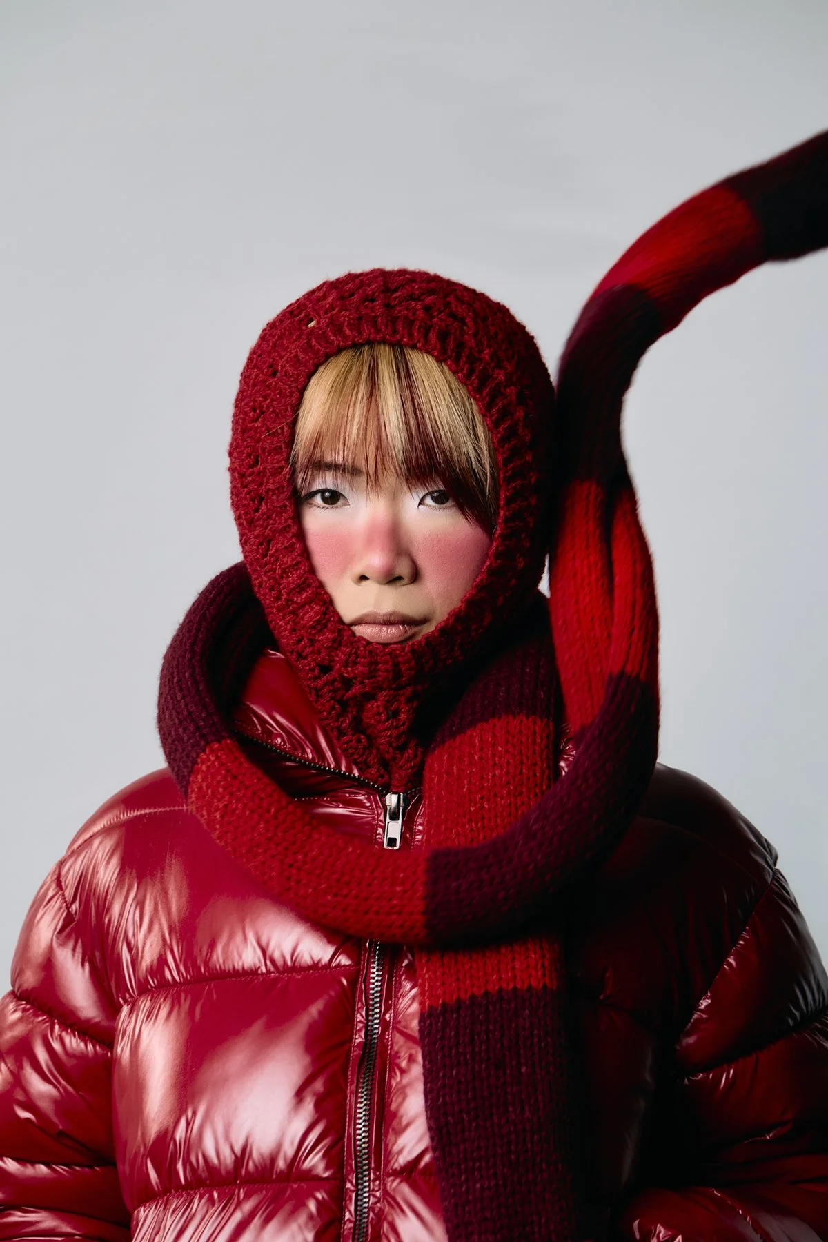 Person wearing a red hooded knit scarf and puffy red jacket, looking directly at the camera against a plain background.