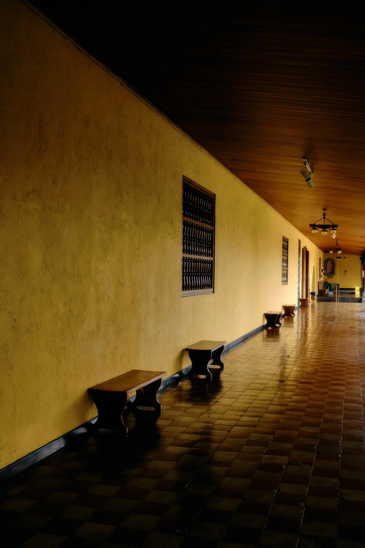 A long, covered walkway with yellow textured walls, wooden benches, and tiled floor, with windows and chandeliers overhead.