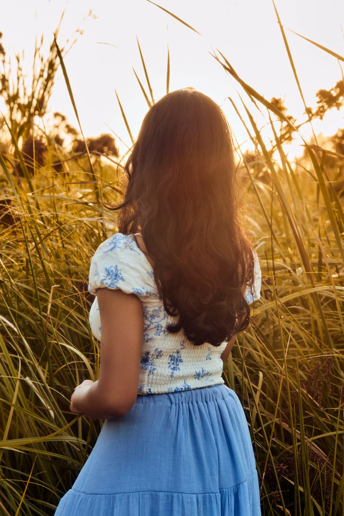 A woman with long brown hair wearing a white and blue top and a blue skirt, standing in tall grass during sunset.