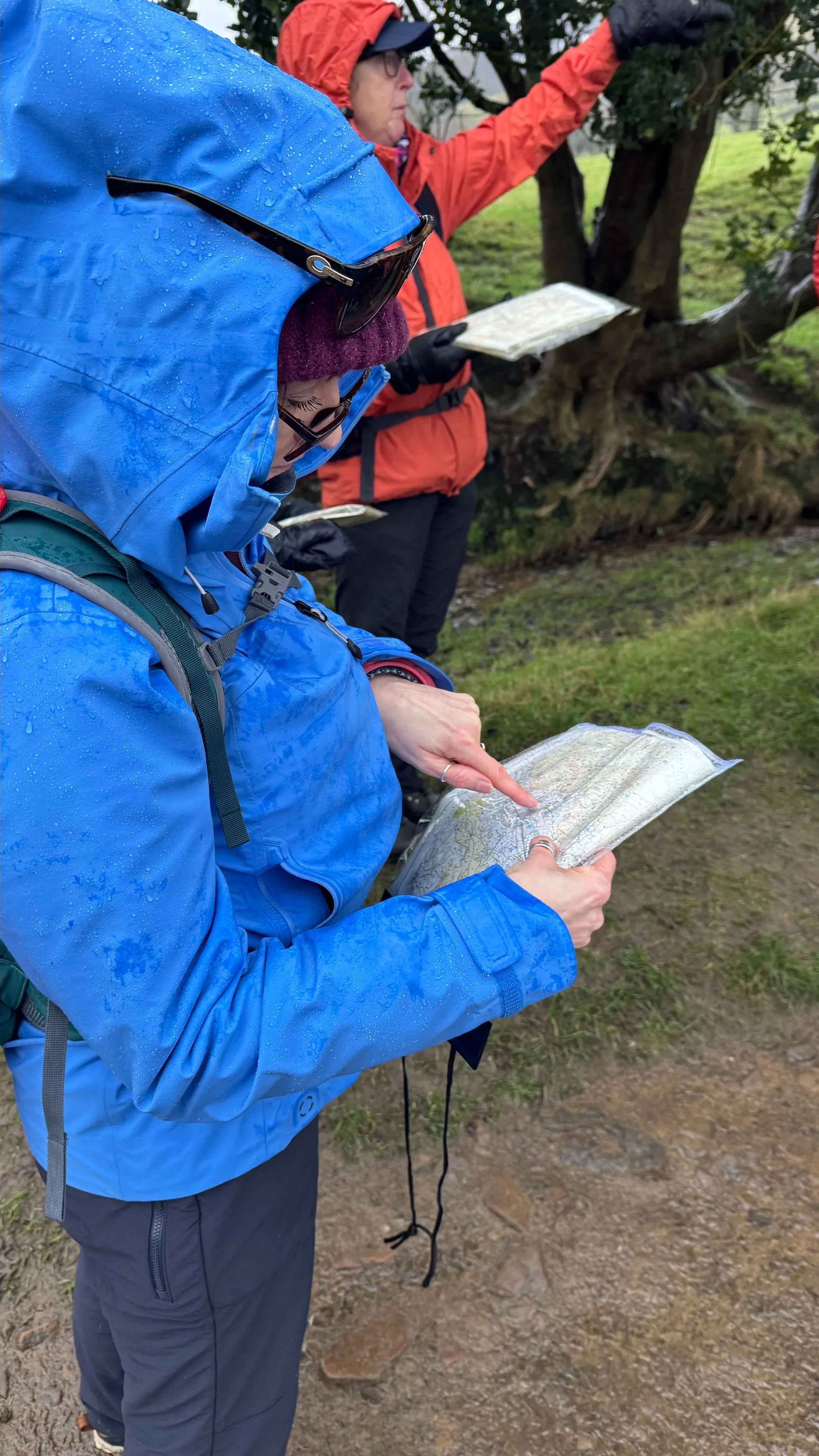 Two people outdoors, dressed in rain jackets, looking at a map. One person is pointing to the map, while the other is observing. The background includes trees and grass, suggesting a natural setting.