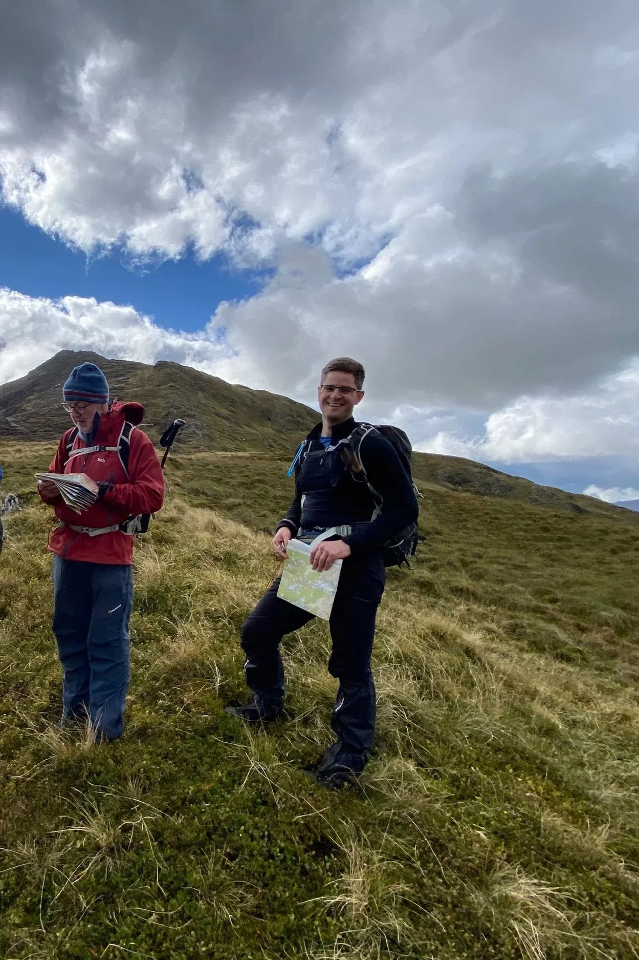 Two hikers on a grassy hillside with mountainous terrain and partly cloudy sky.