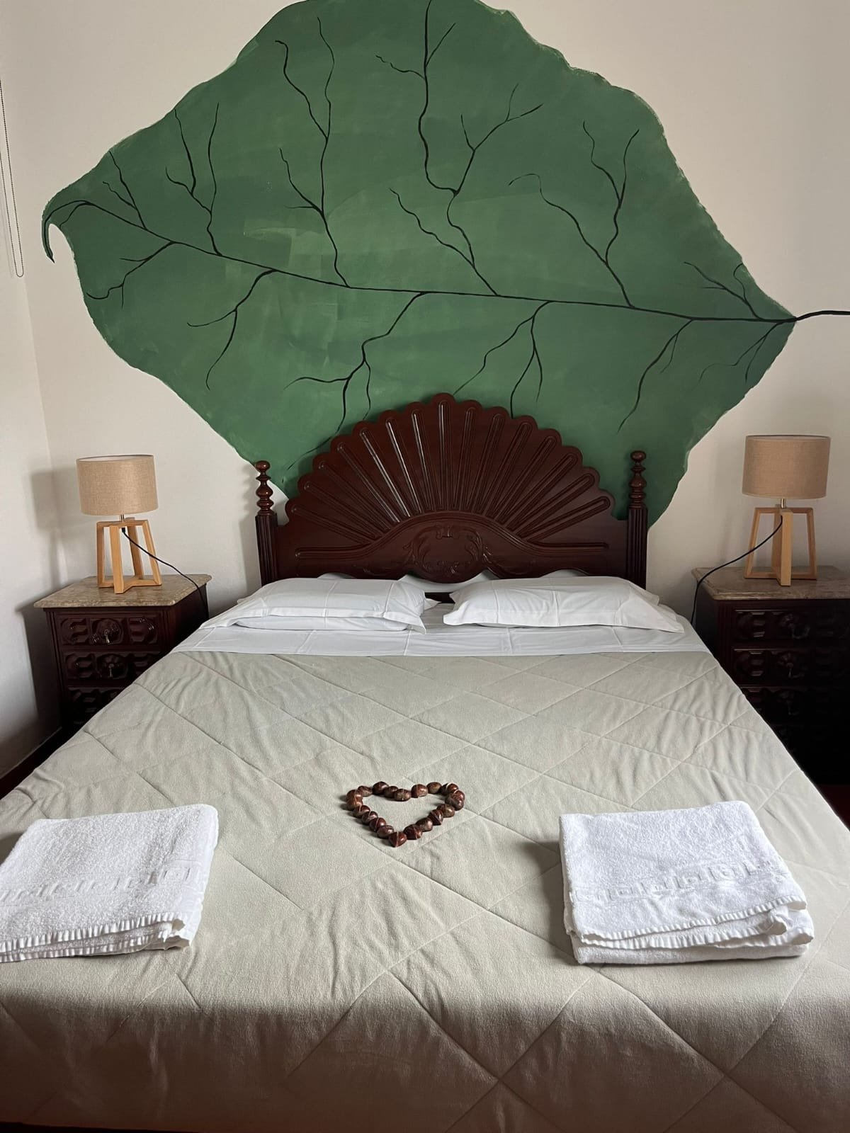 Cama de casal com lençóis brancos, toalhas dobradas na ponta, coração feito de castanhas no centro, cabeceira de madeira com detalhes trabalhados, mural de parede com uma grande folha verde, dois abajures de mesa em ambos os lados da cama.