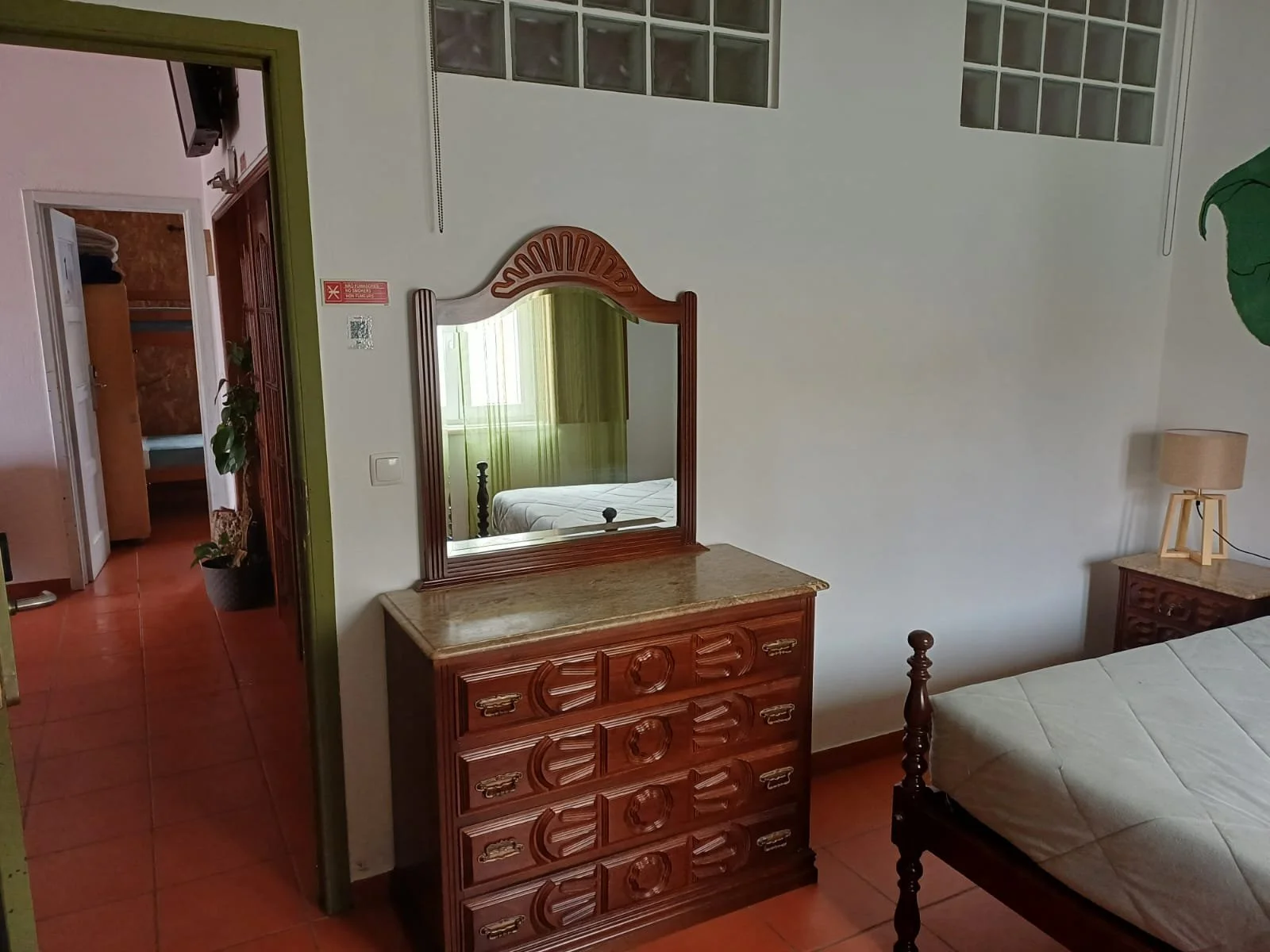 Quarto com cama de madeira, espelho de madeira, cômoda de pedra, lâmpada de mesa, parede branca e piso de azulejo avermelhado.
