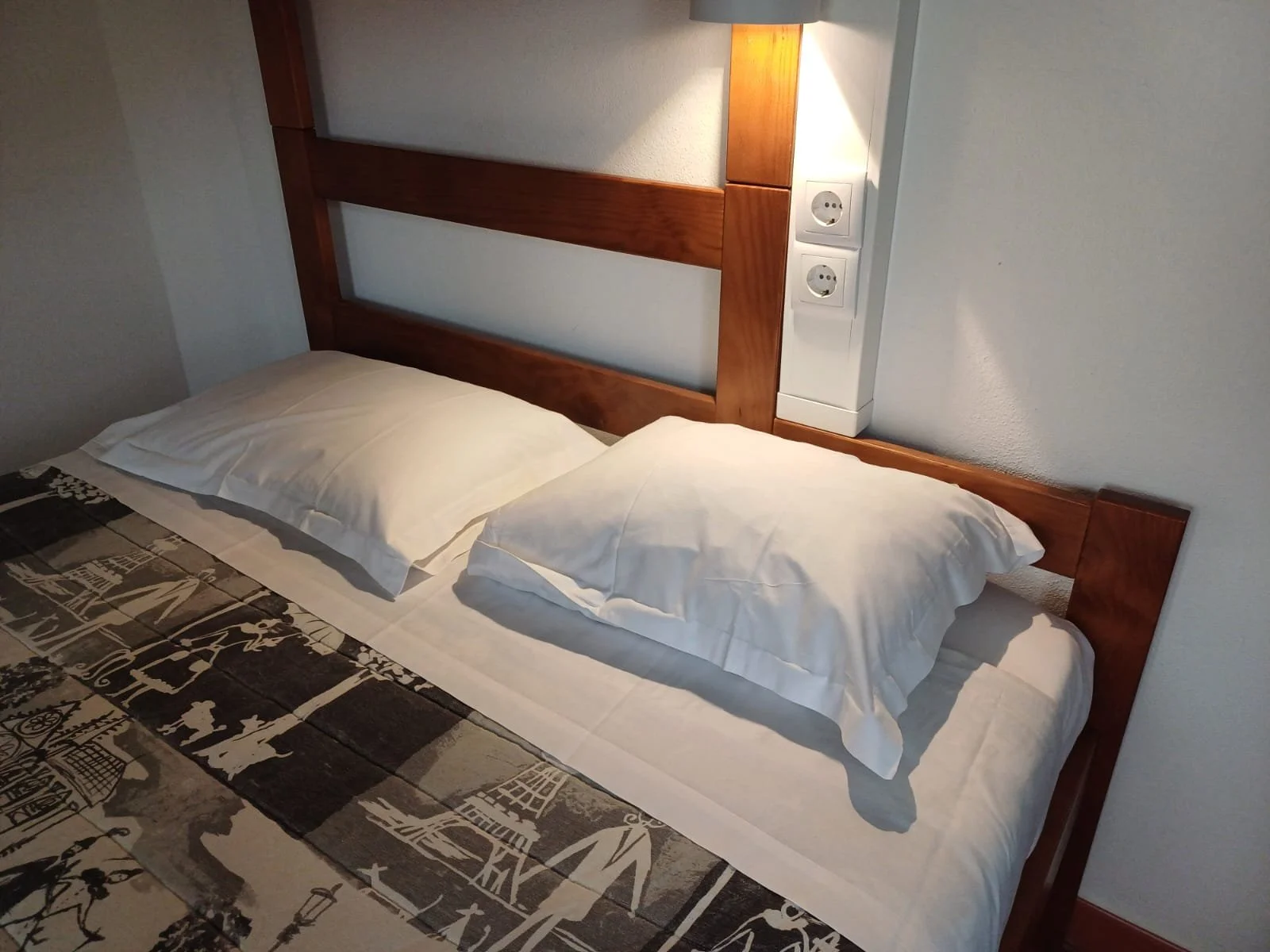 Cama de casal com cabeceira de madeira, travesseiros brancos, roupa de cama branca, edredom com estampa de linhas negras e cinzas, parede branca ao fundo com tomadas elétricas.