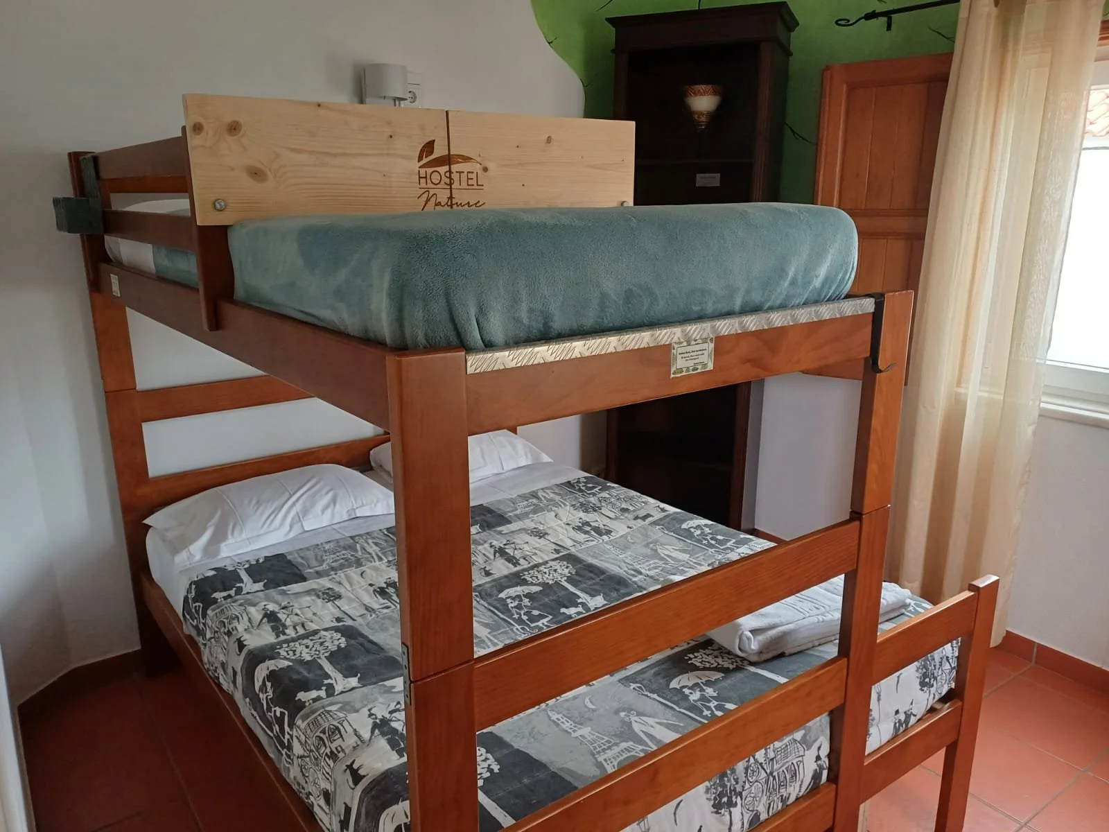 Cama beliche de madeira com colchão de casal na parte inferior e um colchão de solteiro na parte superior, em um quarto com cortinas na janela e móveis de madeira ao fundo.