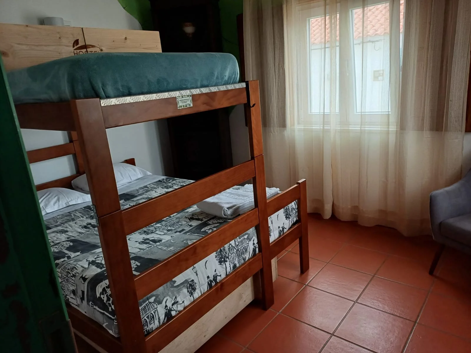 Quarto com beliche de madeira, com lençóis de estampas urbanas, duas camas com travesseiros brancos, janela com cortinas transparentes, cadeira cinza ao lado, piso de azulejos vermelhos.