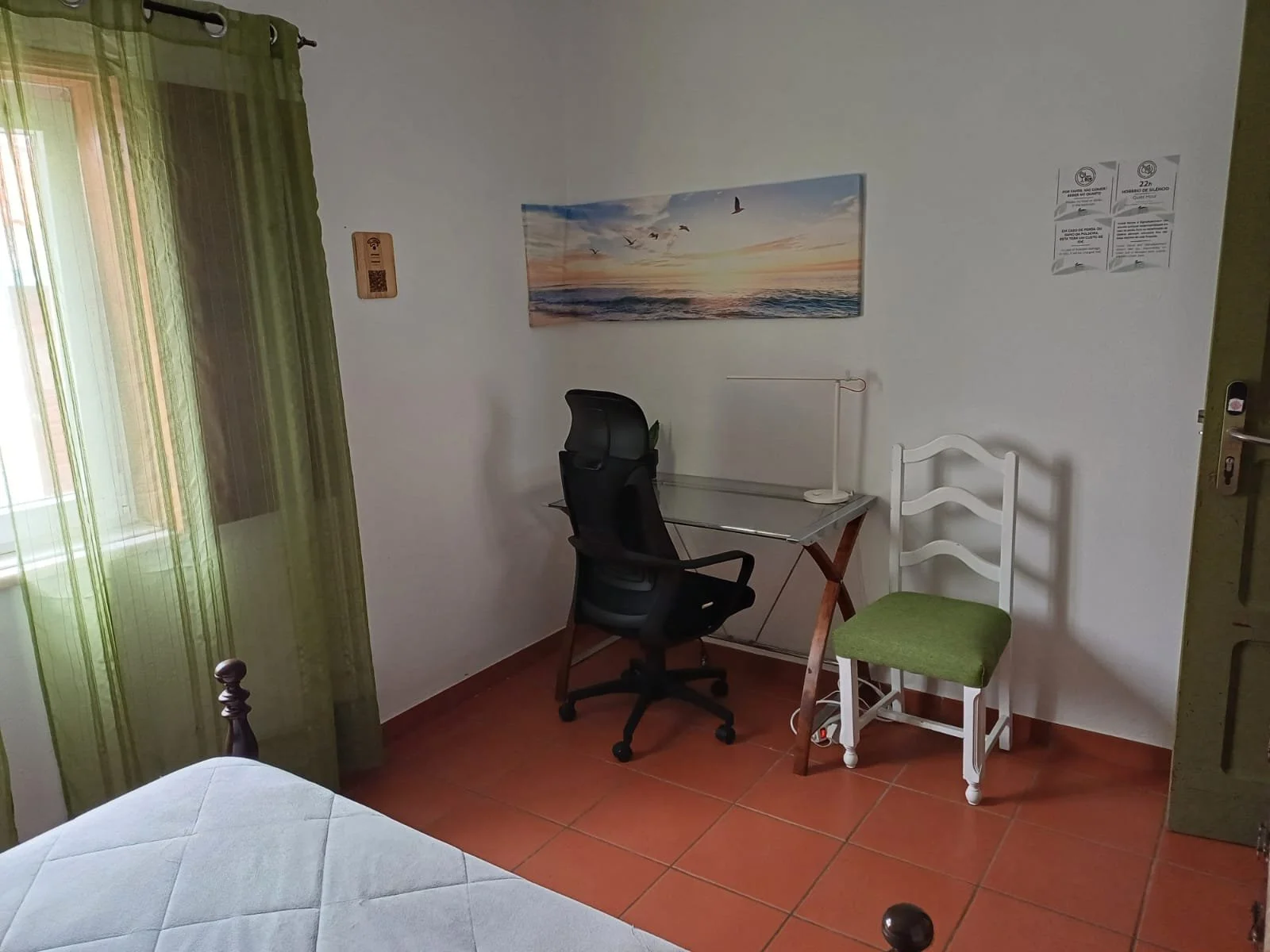 Quarto com janela de cortina verde, uma mesa com cadeira de escritório preta, uma cadeira branca com assento verde, uma pintura de pôr do sol com pássaros no muro e um cartaz de informações na parede