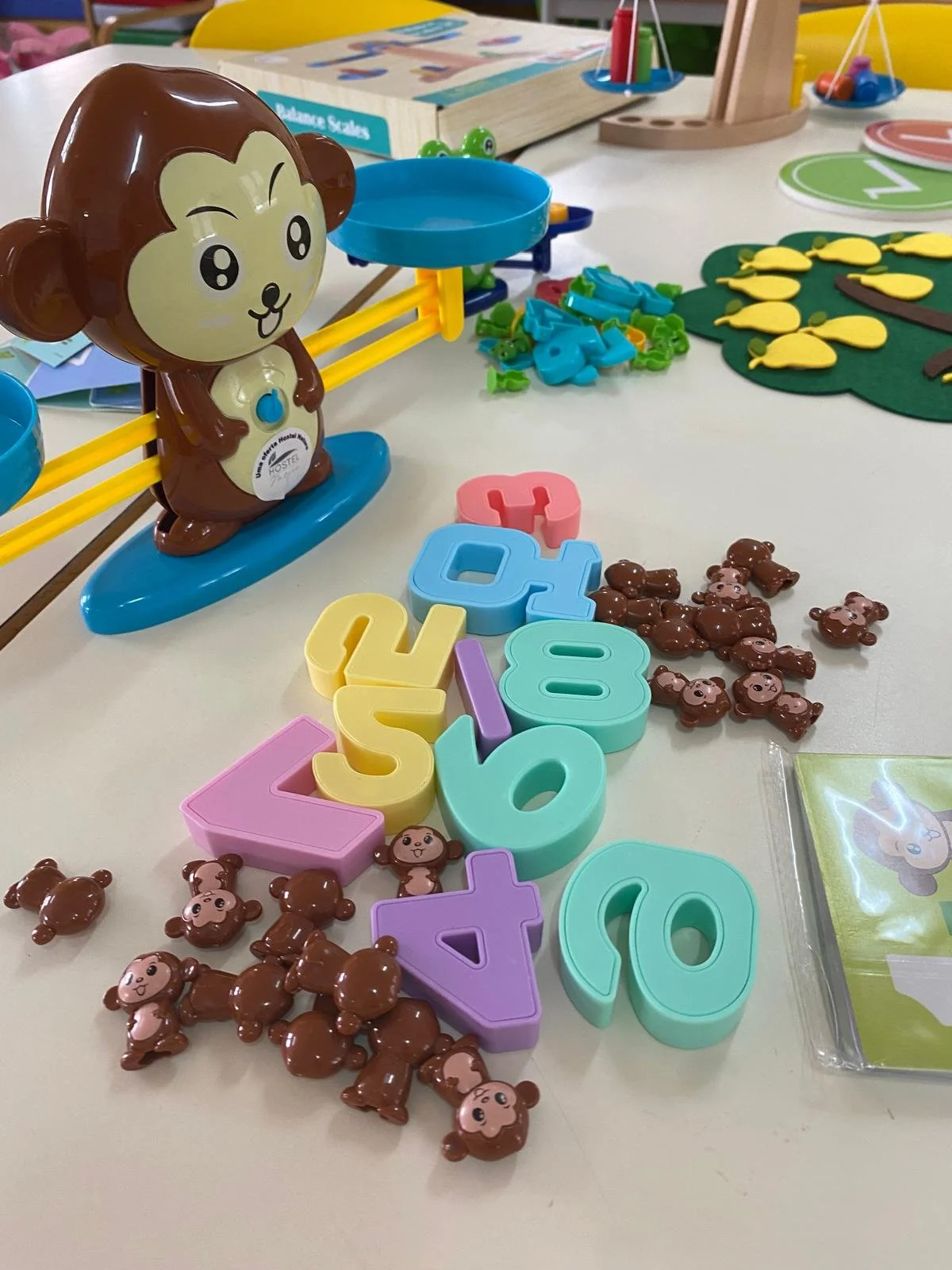 Brinquedos de diferentes cores e formas, incluindo letras, ursinhos de pelúcia de plástico, uma balança de brinquedo e peças de jogo, dispostos sobre uma mesa.