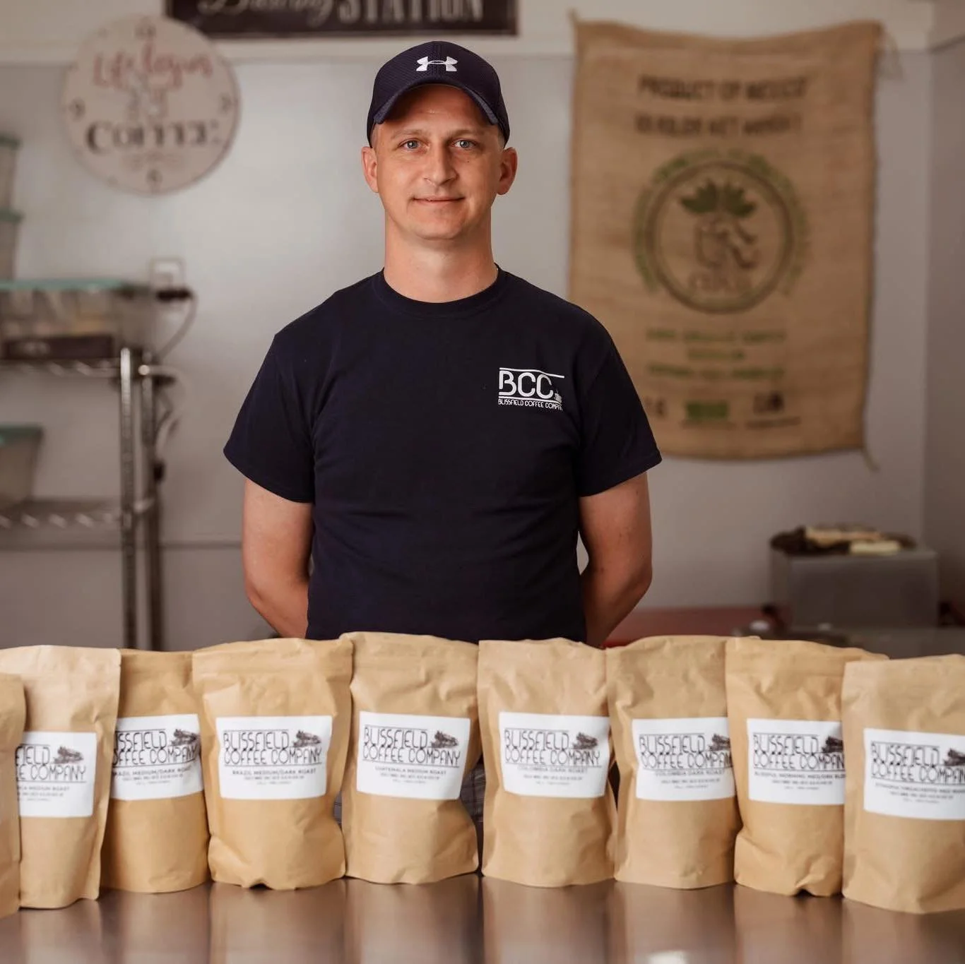 A man in a black t-shirt and black cap standing behind a row of brown paper coffee bags labeled 'Blisfield Coffee Company' inside a coffee shop or roastery.