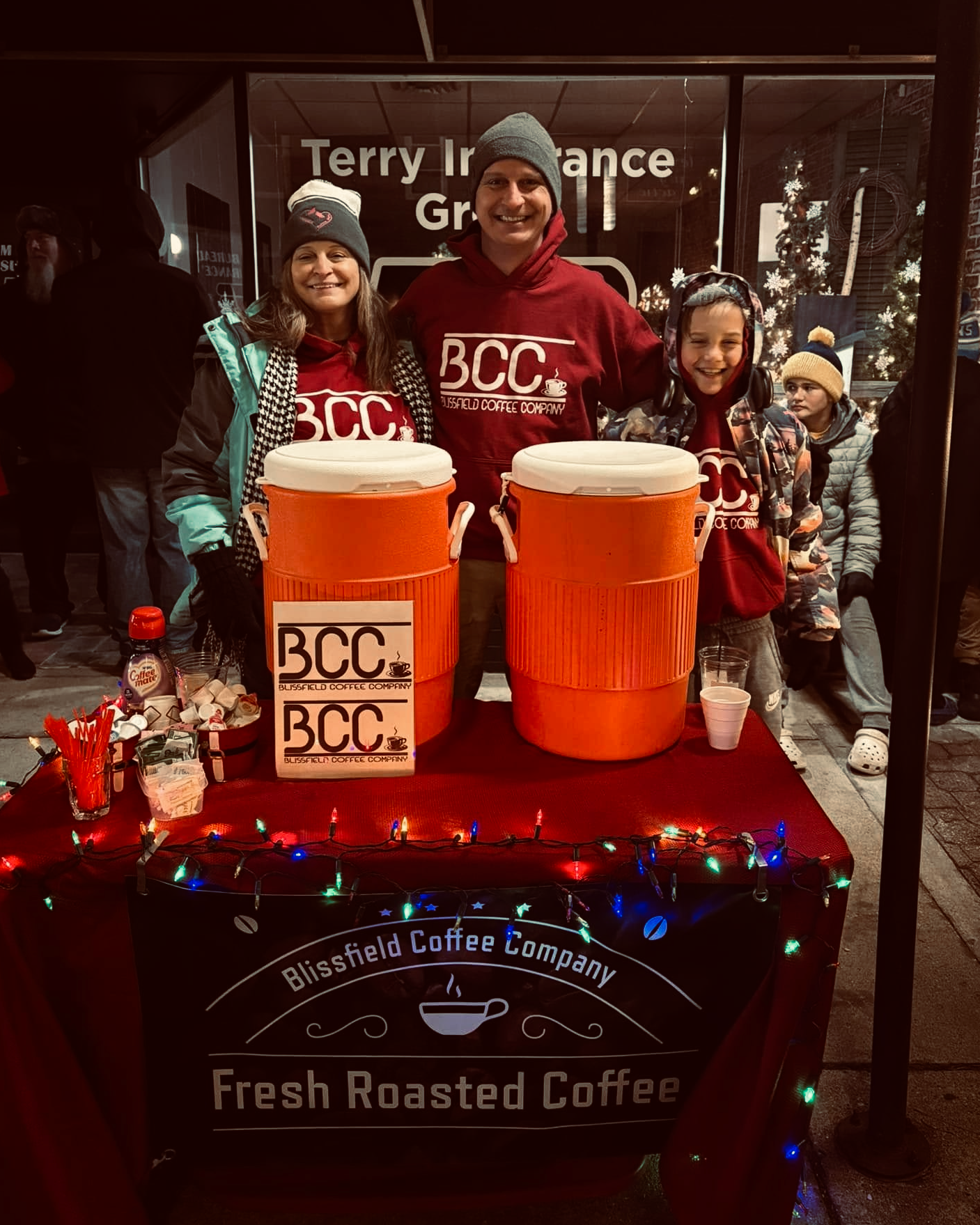 People smiling behind a table with 2 large orange dispensers labeled BCC, surrounded by Christmas decorations and lights, in front of a downtown storefront.
