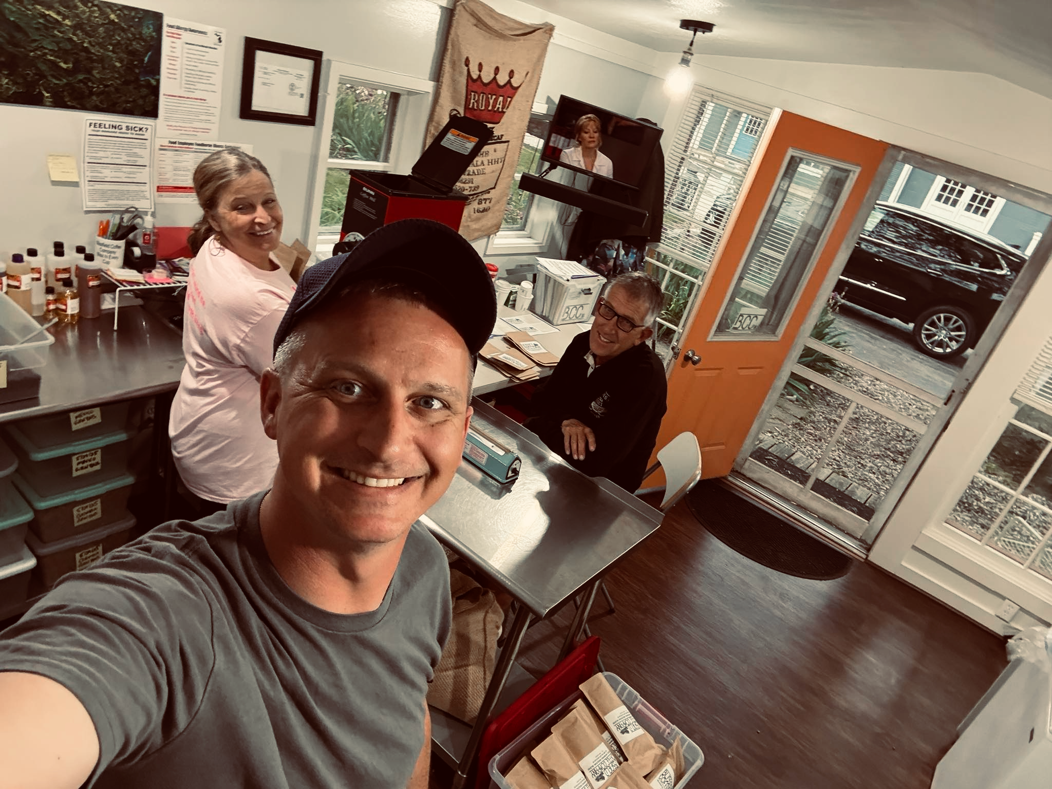A man in a gray t-shirt taking a selfie in a room with several people. Behind him, a woman smiling at the camera is sitting at a table with various bottles and containers. Two other people, a man and a woman, are sitting at the table. The room has large windows, orange and white walls, and various posters and a banner hanging on the wall.