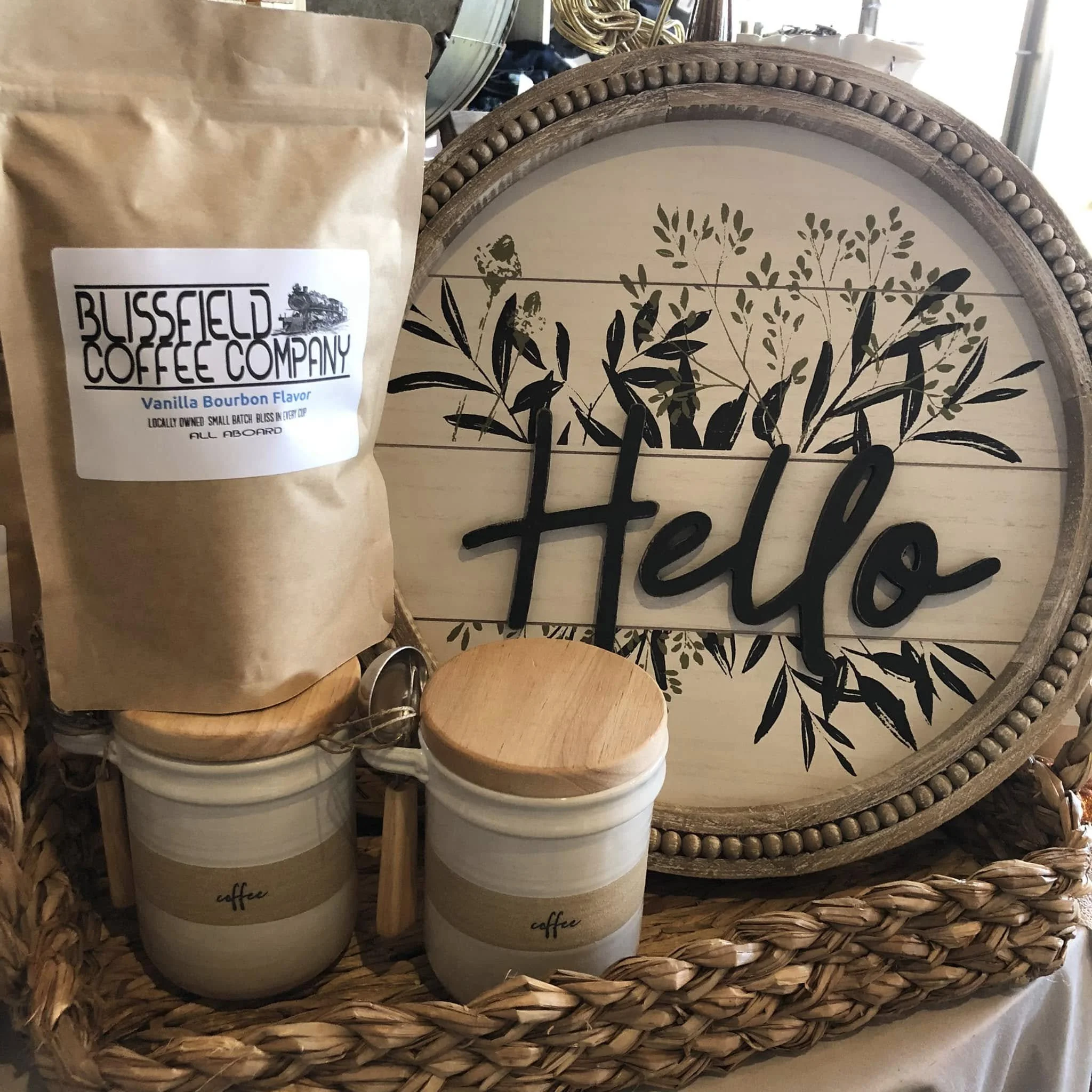 A paper coffee bag labeled 'Blissfield Coffee Company' with vanilla bourbon flavor, placed on top of white candles with wooden lids that have 'coffee' written on them. The background features decorative home decor and plants on a wicker tray.