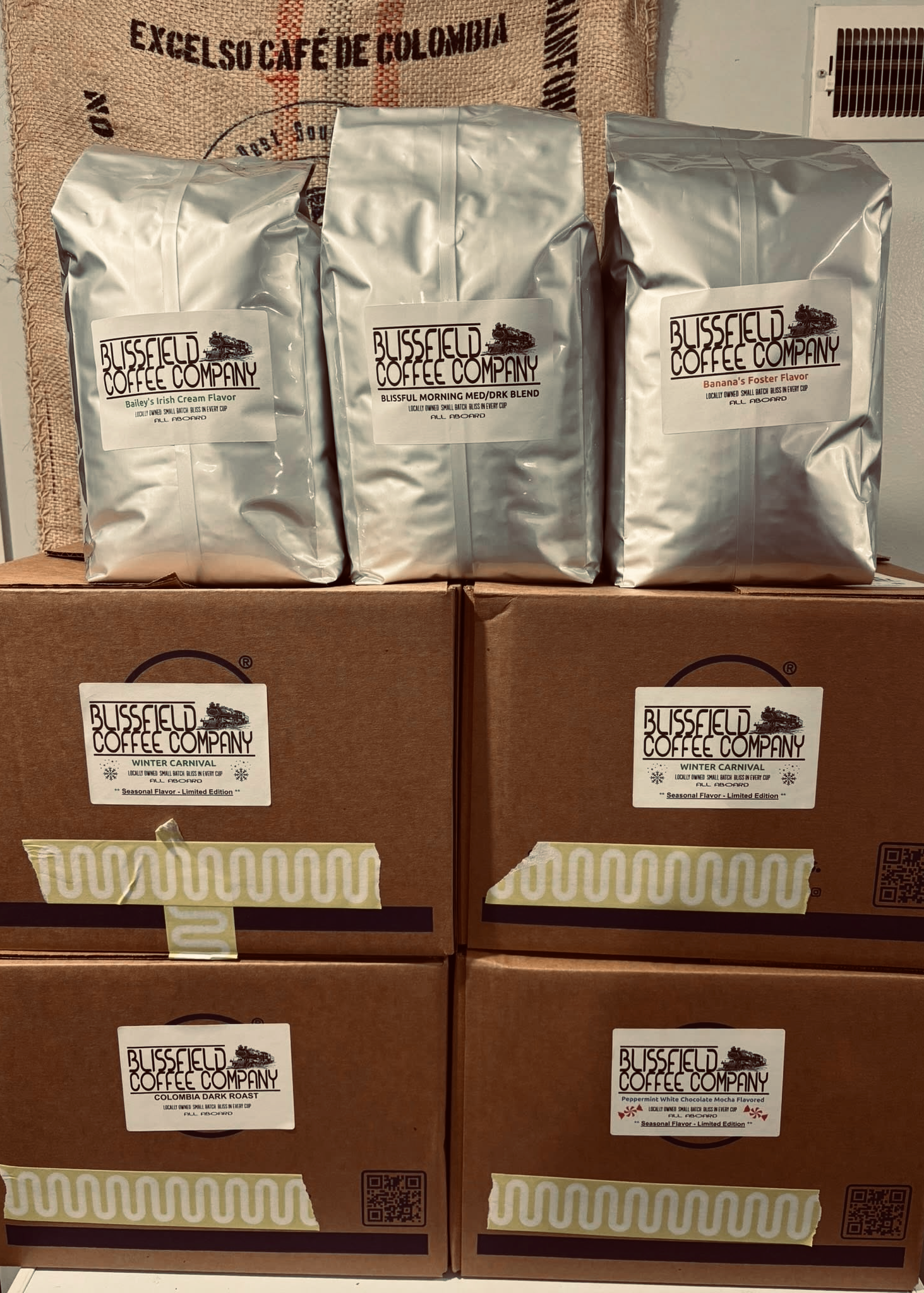 Three bags of coffee from Blusfield Coffee Company, labeled Bailey's Irish Cream Flavor, Blissful Morning Med/Drk Blend, and Banana's Foster Flavor, placed on a cardboard box with a burlap coffee sack in the background.