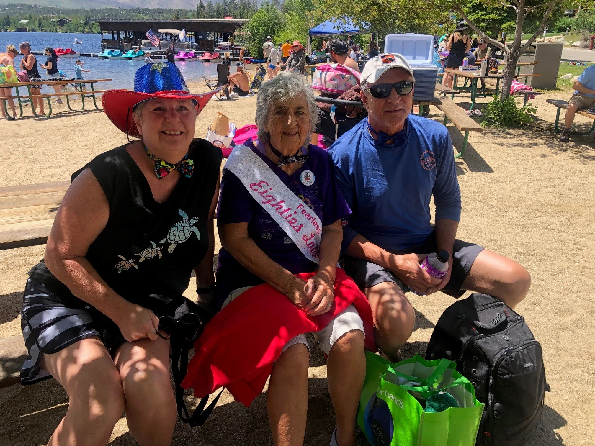 Three people sitting on a park bench at a lakeside event, smiling and dressed in summer clothes, with water activities and tents in the background.