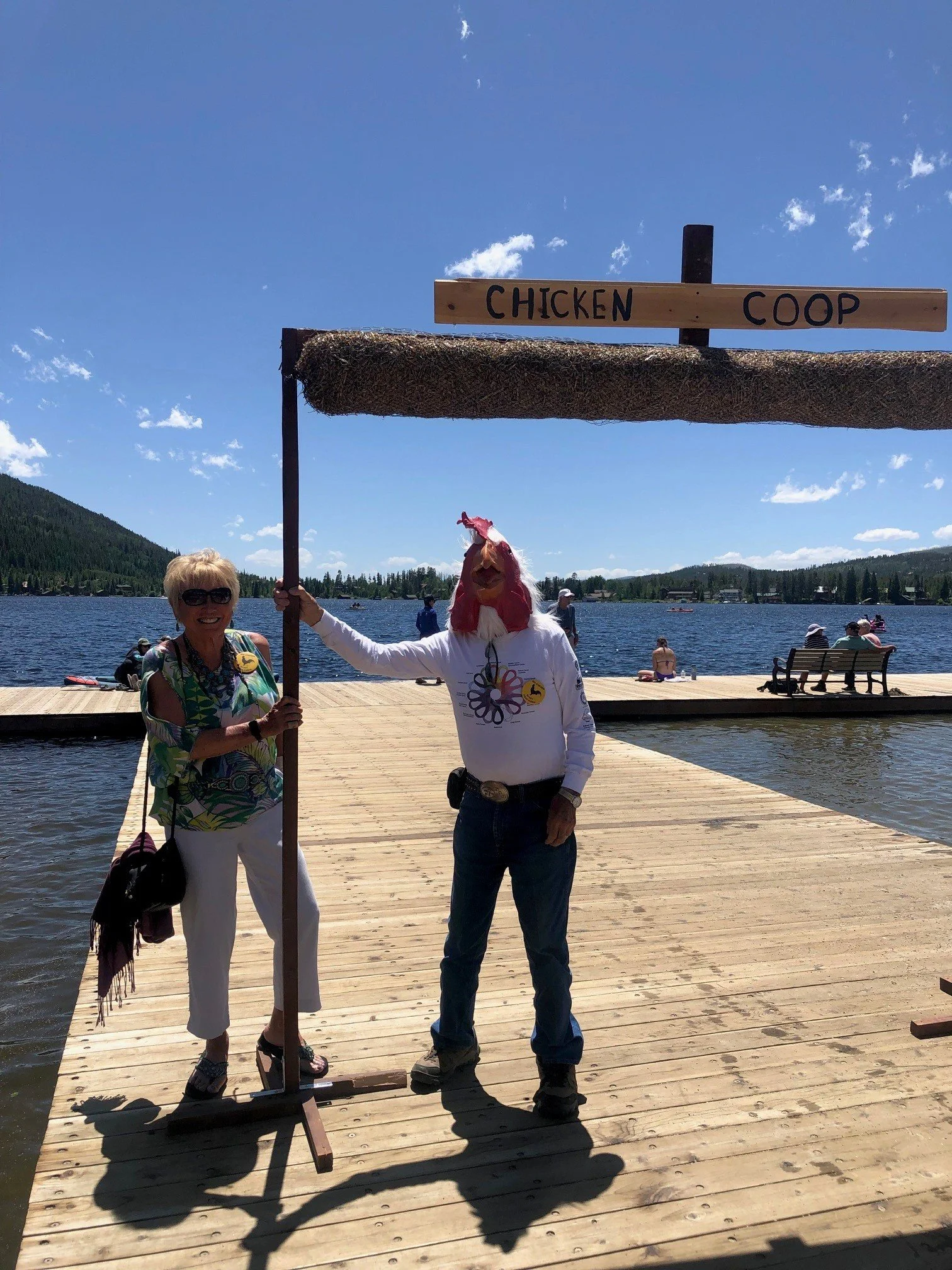 Two people standing on a wooden dock by a lake, one dressed as a chicken, holding a sign that reads 'Chicken Coop'. The sky is clear with some clouds, and there are a few people in the background near the water.