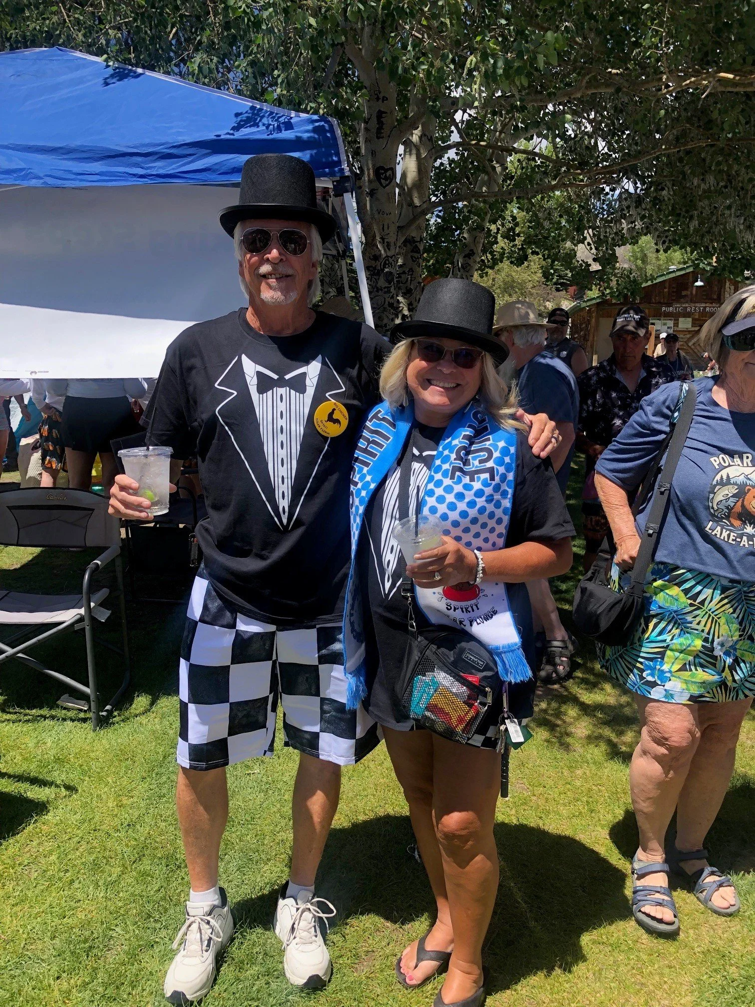 A man and woman standing outdoors at a festival or outdoor event, both wearing black top hats, sunglasses, and themed black shirts. The man is dressed in a tuxedo-themed shirt with checkered shorts, holding a drink, and the woman is wearing a scarf,