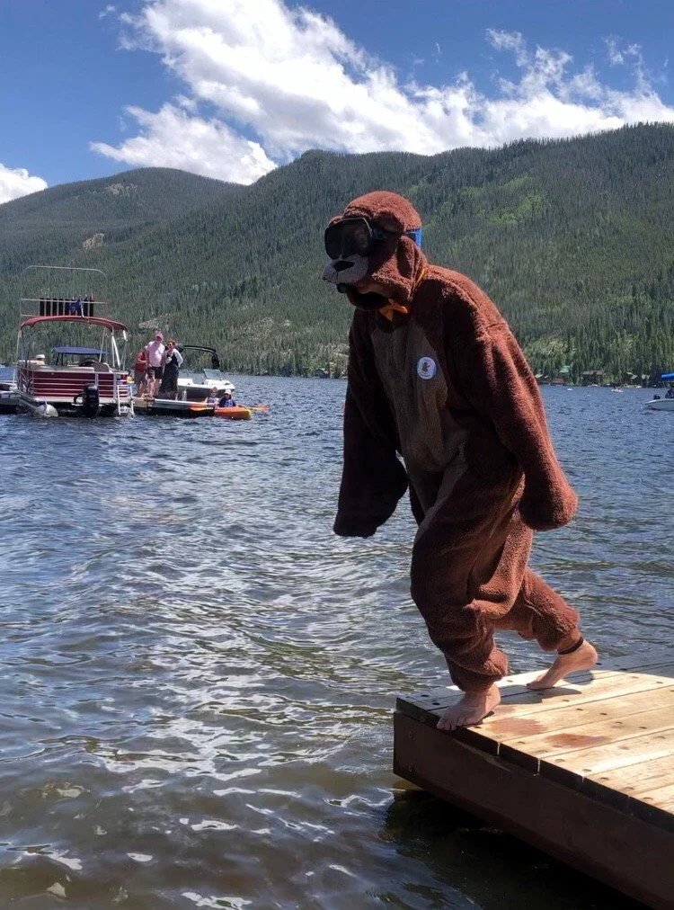 Person in a bear costume with sunglasses standing at the edge of a dock by a lake, with boats and forested mountains in the background.