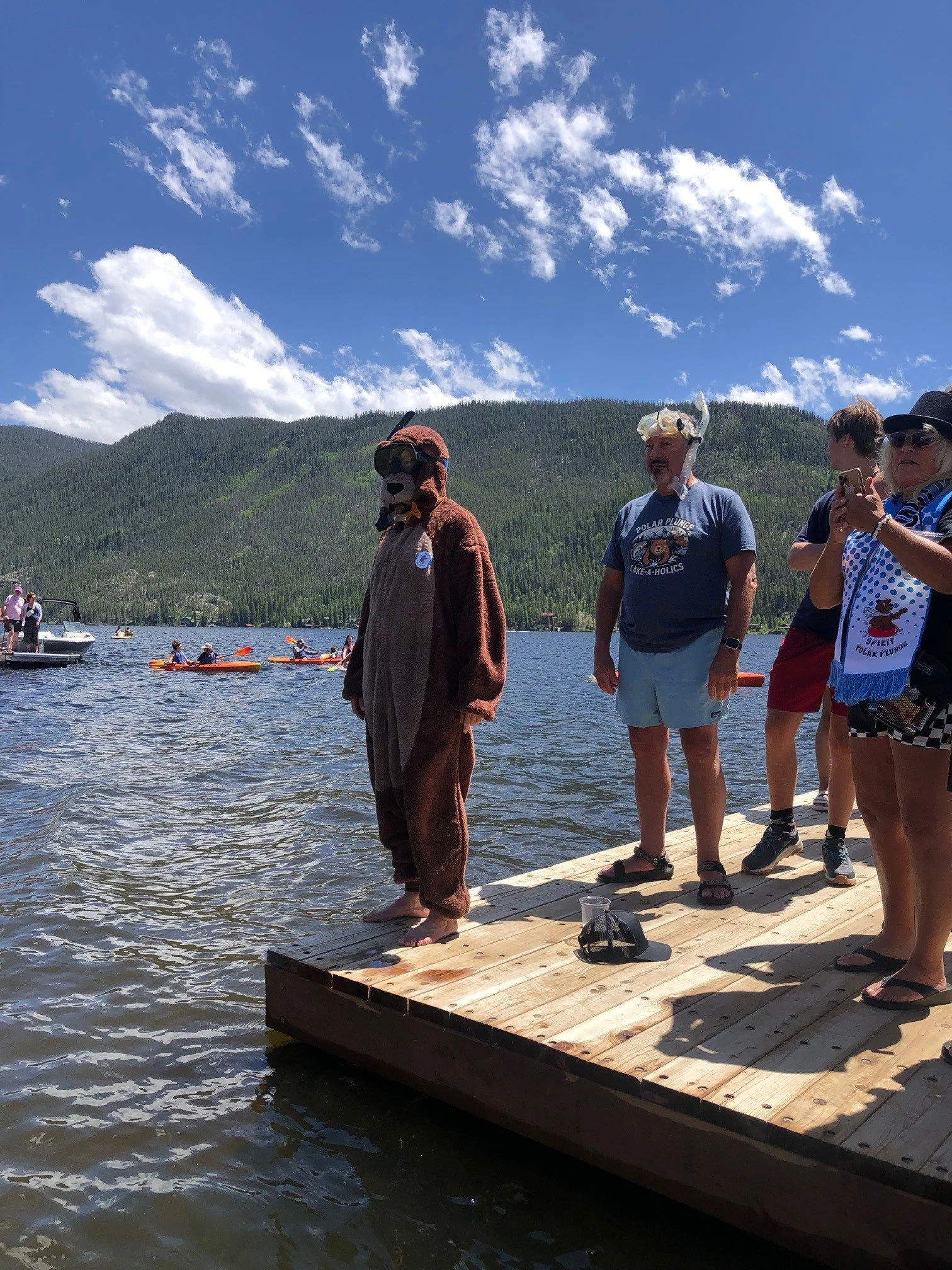 People standing on a wooden dock at a lake, with a person dressed in a bear costume. In the background, there are kayak paddlers, boats, a forested mountain, and a partly cloudy sky.