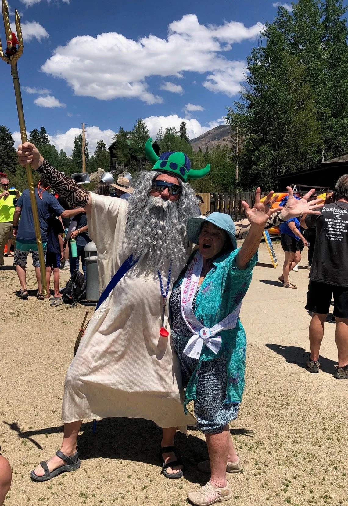 Two people dressed in costumes celebrating outdoors on a sunny day. One is wearing a Viking helmet, sunglasses, a long gray beard, and a beige dress, holding a staff. The other is wearing a blue sunhat, a green patterned shirt, and has a sash around