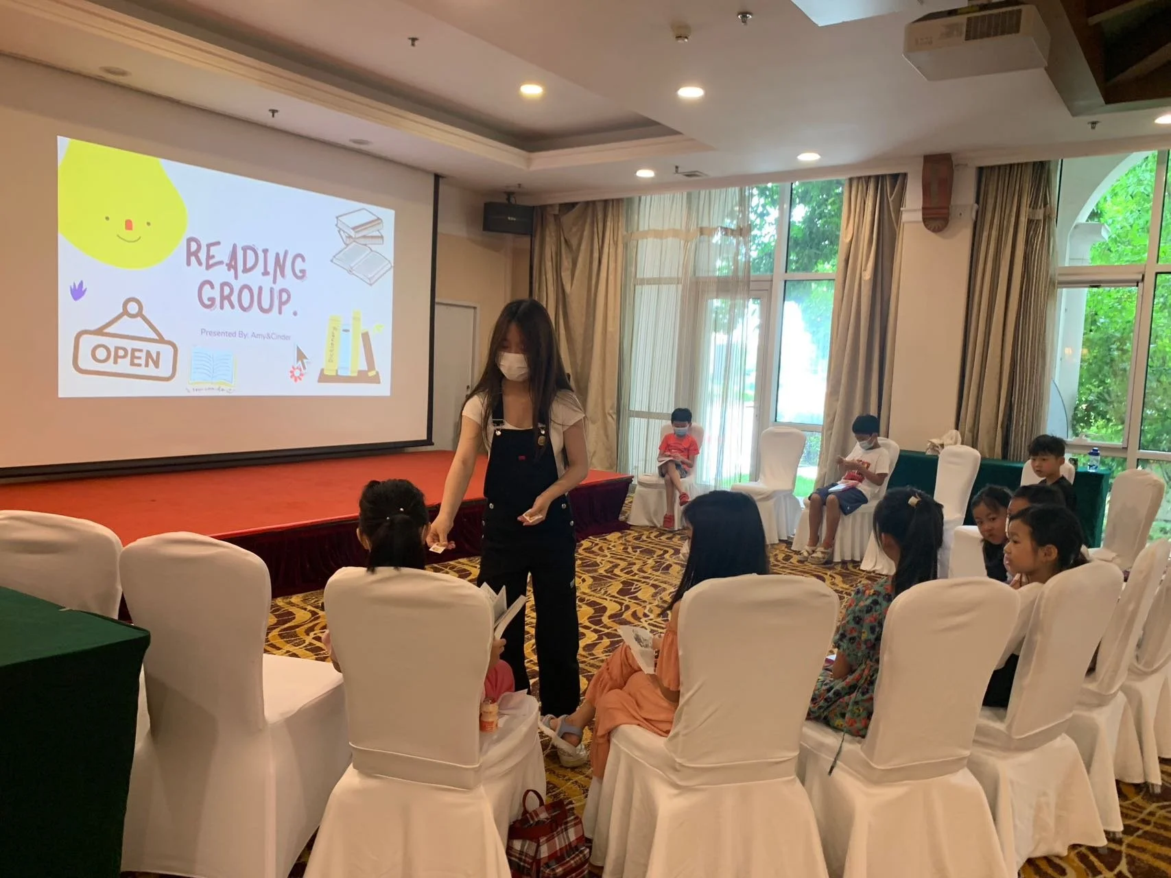 A group of children sitting in white chairs inside a classroom or event room, listening to a woman in front who is holding a booklet. The woman is wearing a face mask. There is a large screen displaying a colorful slide titled 'Reading Group,' with i