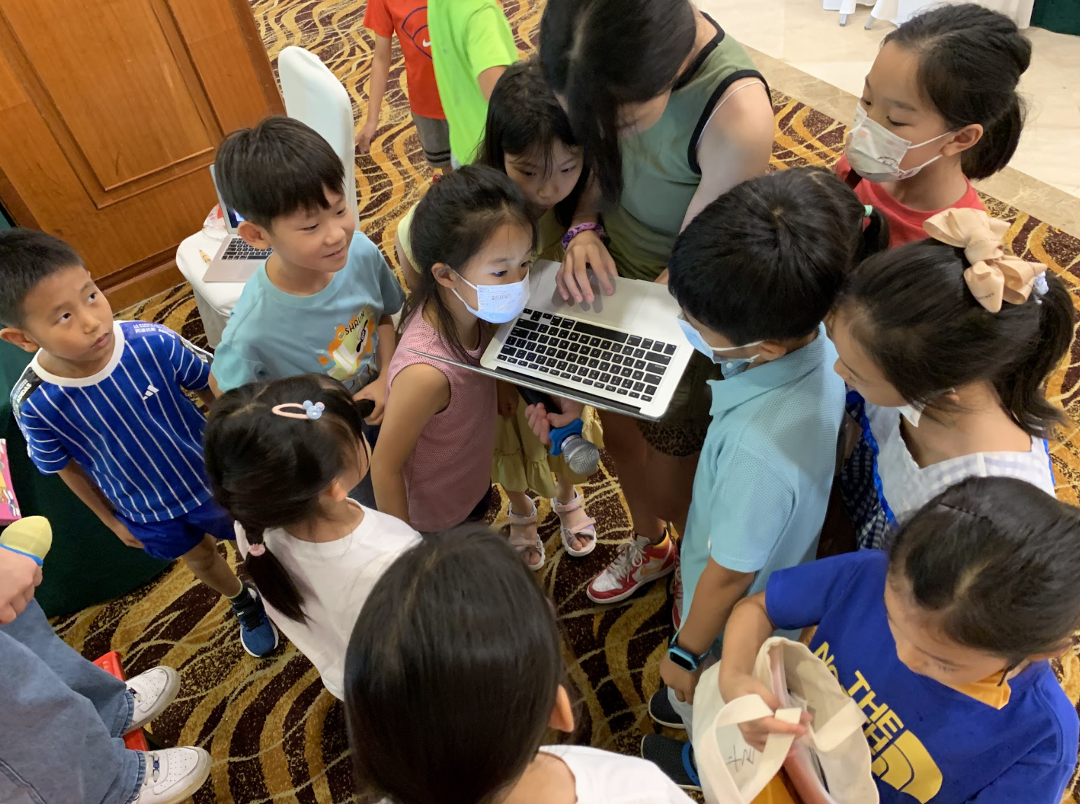Group of children and an adult gathered around a laptop, all wearing face masks, in an indoor setting with carpeted floor.