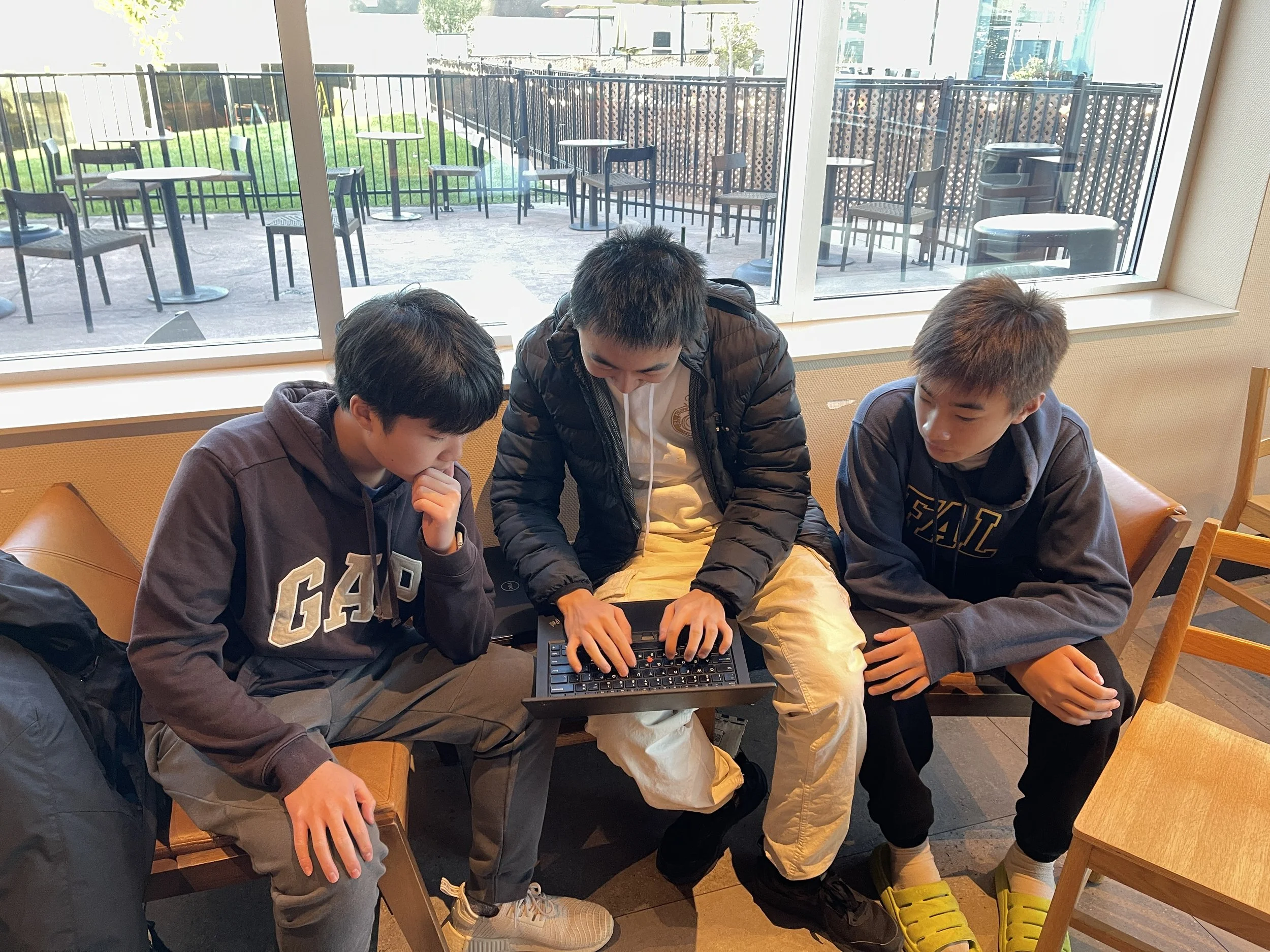 Three boys sitting on chairs indoors, focused on a small keyboard-like device, with large window showing an outdoor patio with tables and chairs.