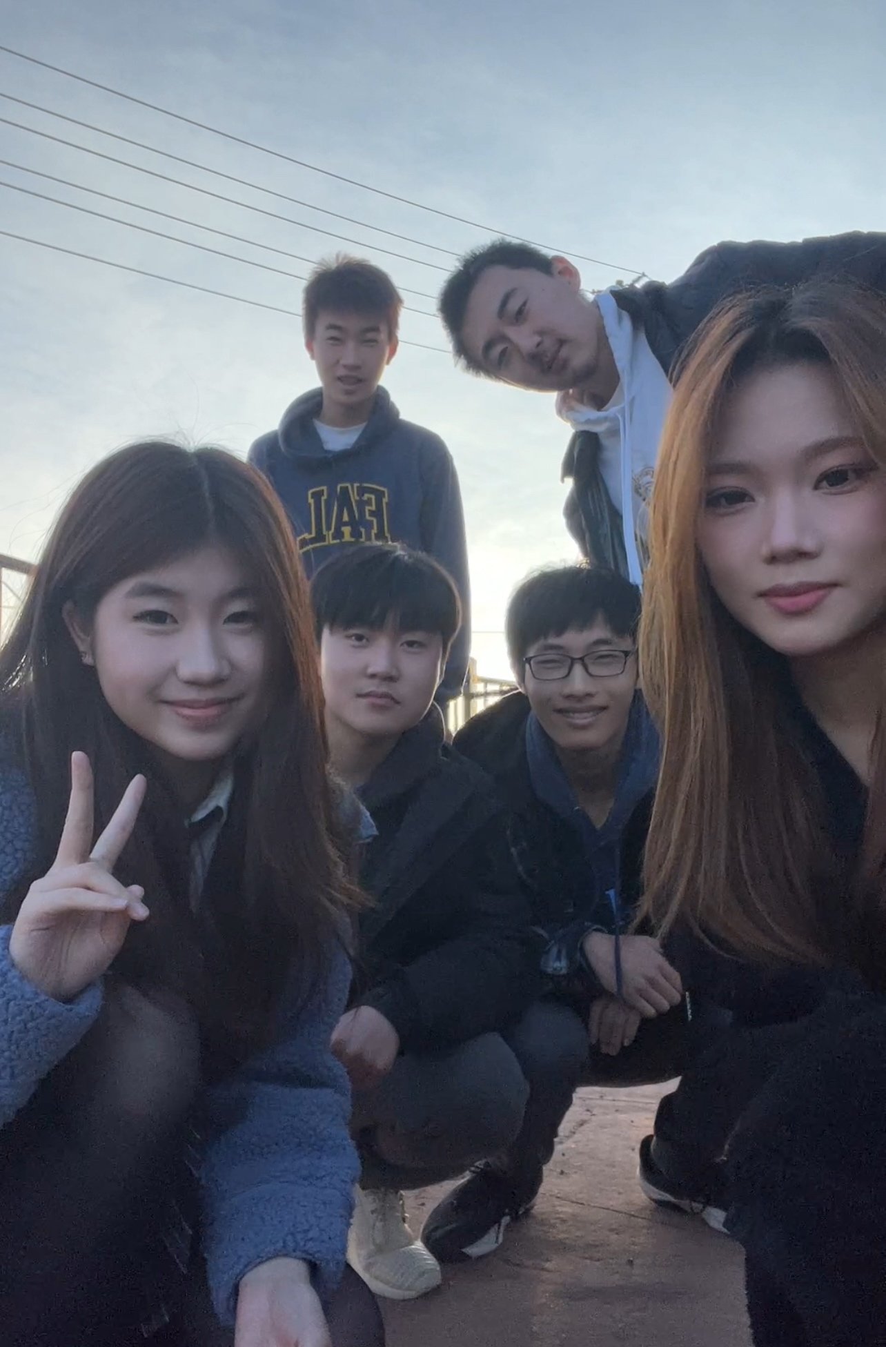 Group of six young people posing outdoors during late afternoon or early evening with a clear sky and power lines in the background.