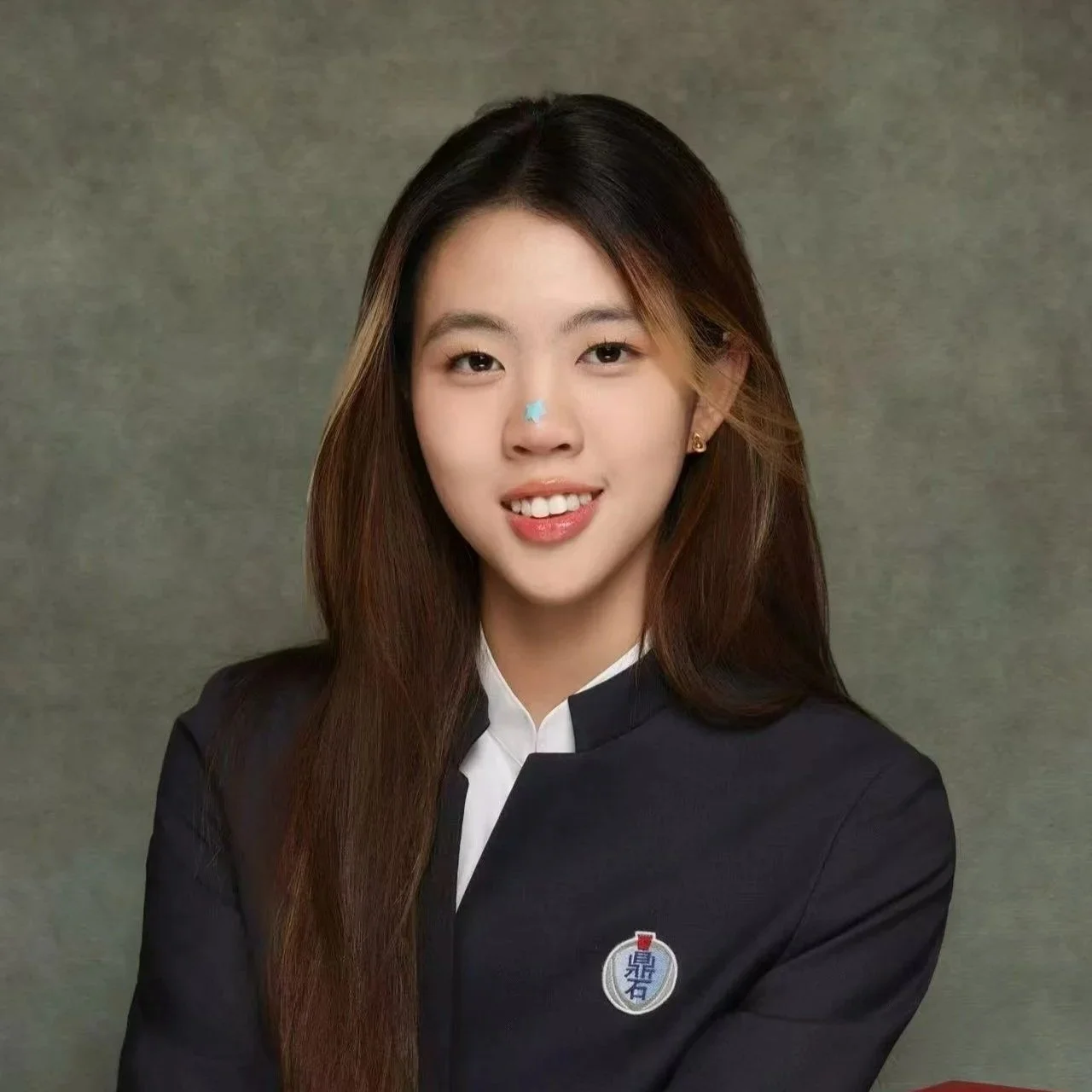 Portrait of a young Asian woman with long brown hair, smiling, wearing a black blazer with a white shirt and a pin on her lapel, against a plain background.
