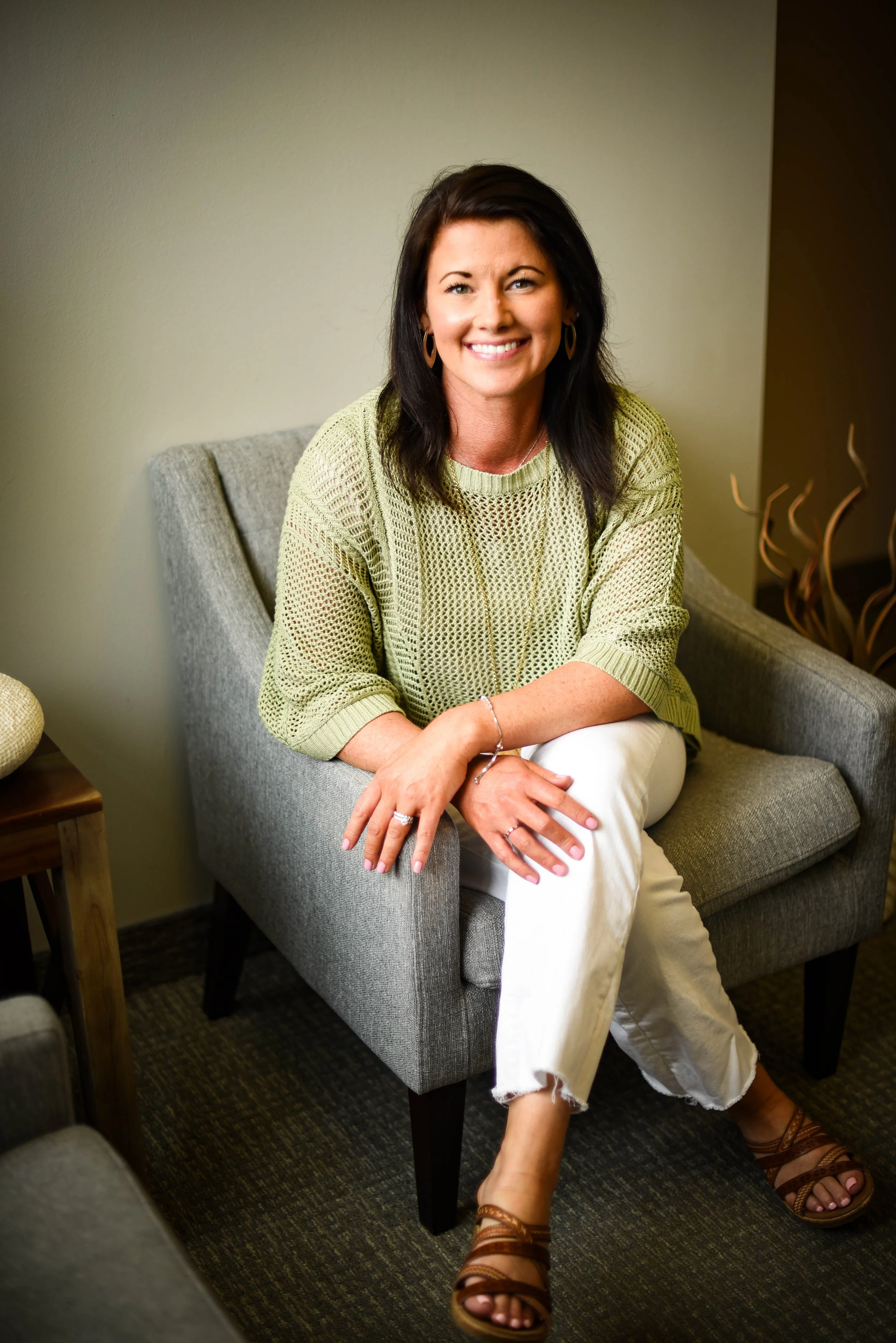 A smiling woman with dark hair wearing a light green knit sweater, white pants, and brown sandals, sitting on a gray armchair.