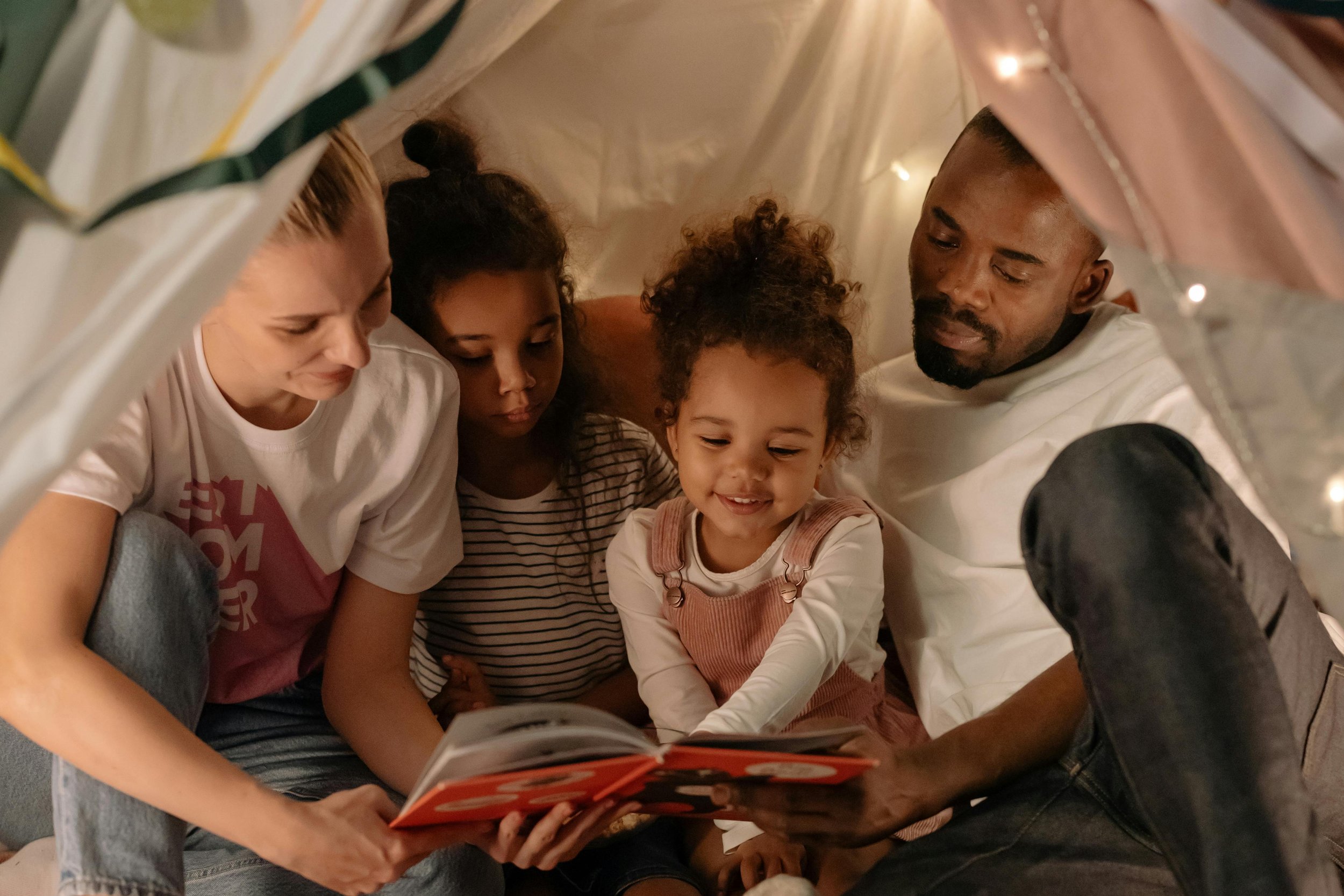 Family of five sitting inside a blanket fort, reading a book together during a cozy moment.
