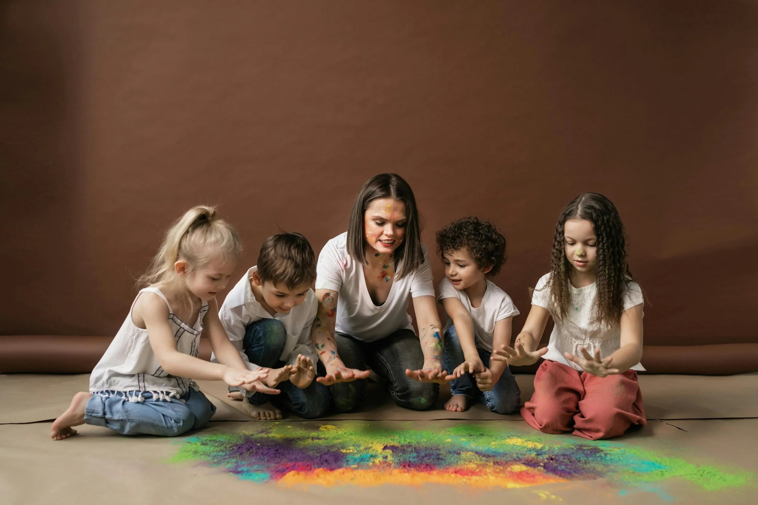 A woman and four children sitting on the floor, playing with colorful powder.