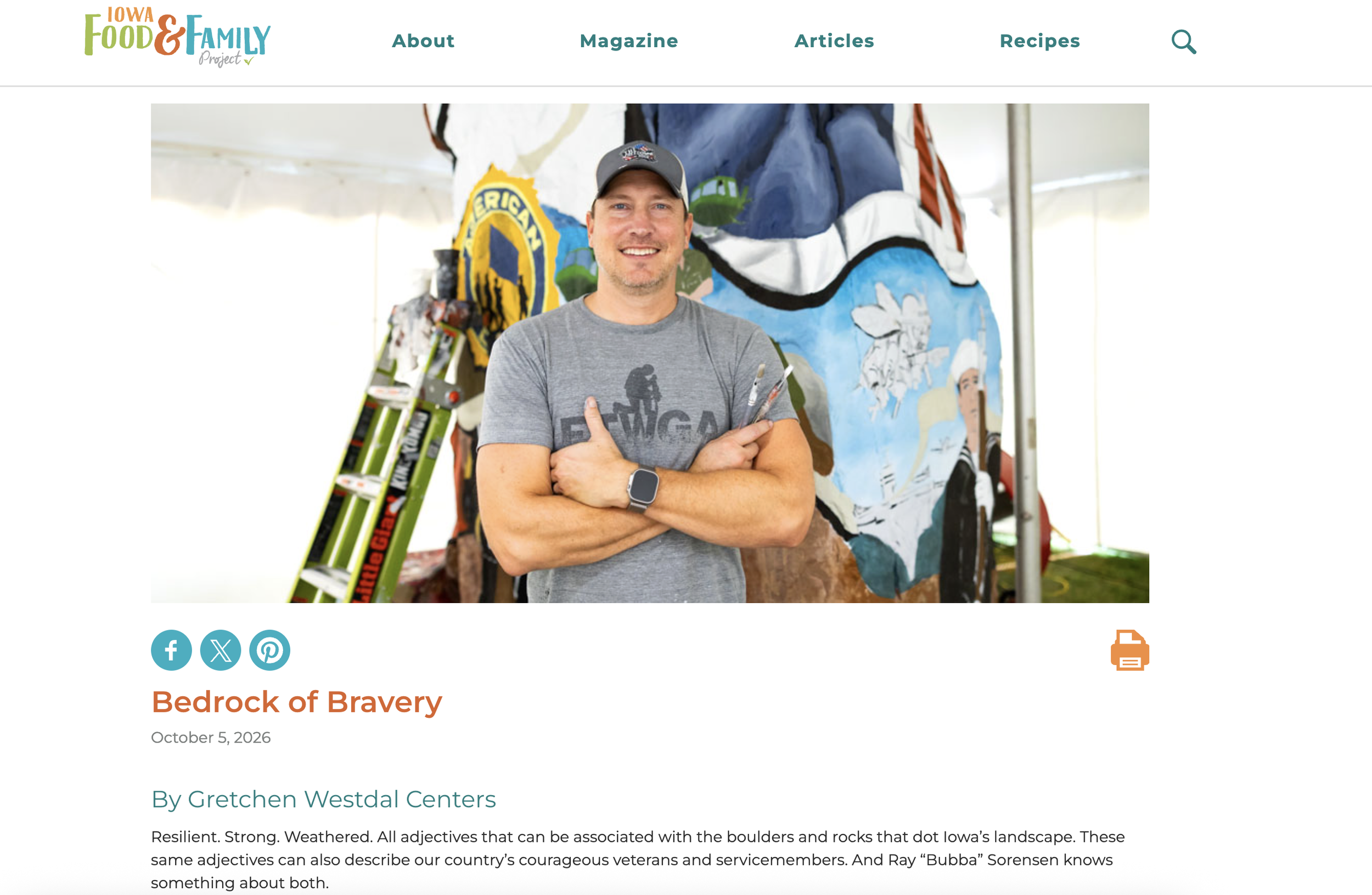 A smiling man wearing a gray t-shirt and baseball cap standing in front of a painted rock with a mural. The mural depicts a person skiing, mountains, forests, and the word 'Erica.'
